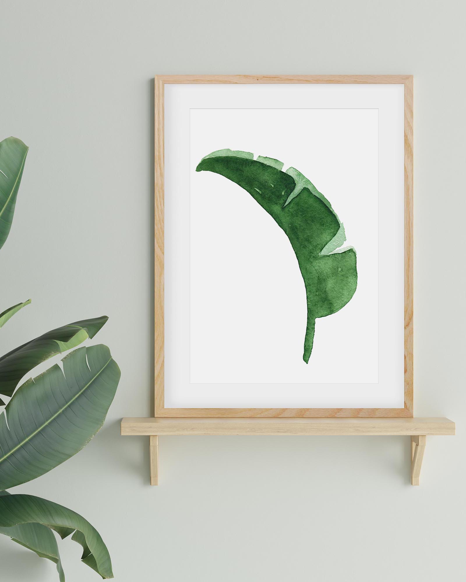 Framed artwork of a green leaf on a white background, leaning against a light gray wall with a plant to the left.