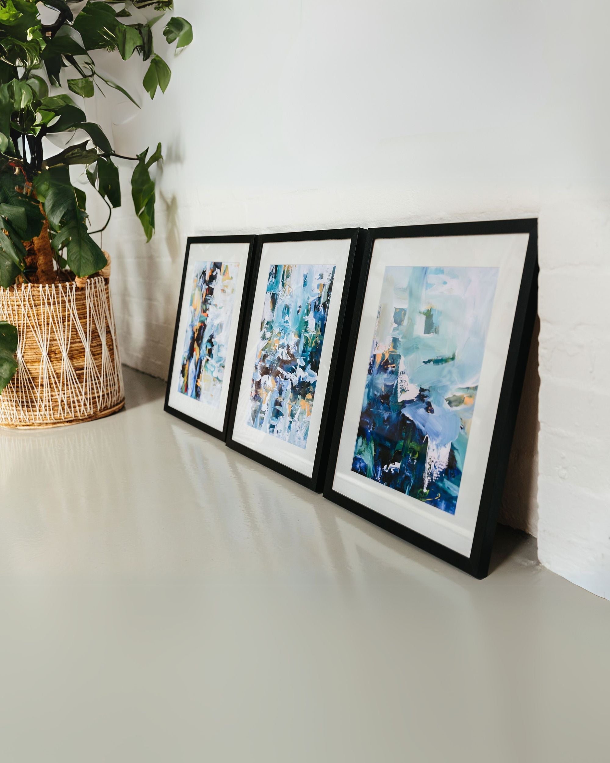 Three framed abstract paintings on a white surface with a plant in a wicker basket.