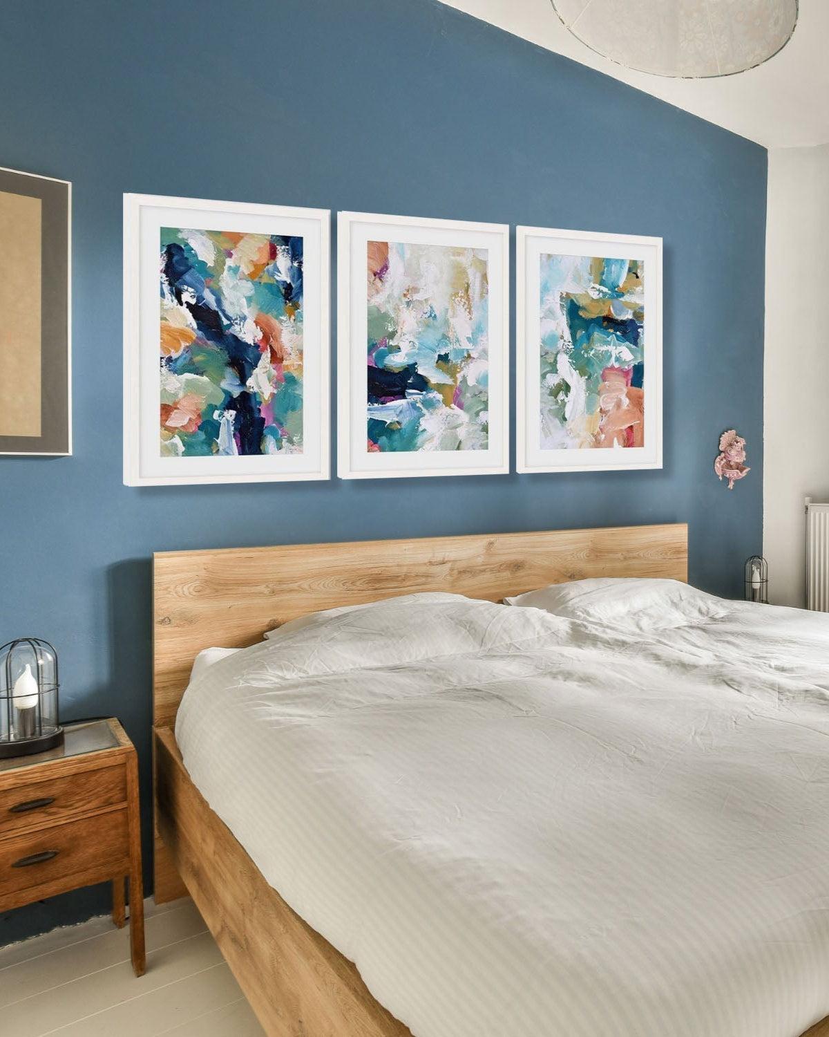 A set of three framed abstract art prints hanging above a bed on a blue wall.