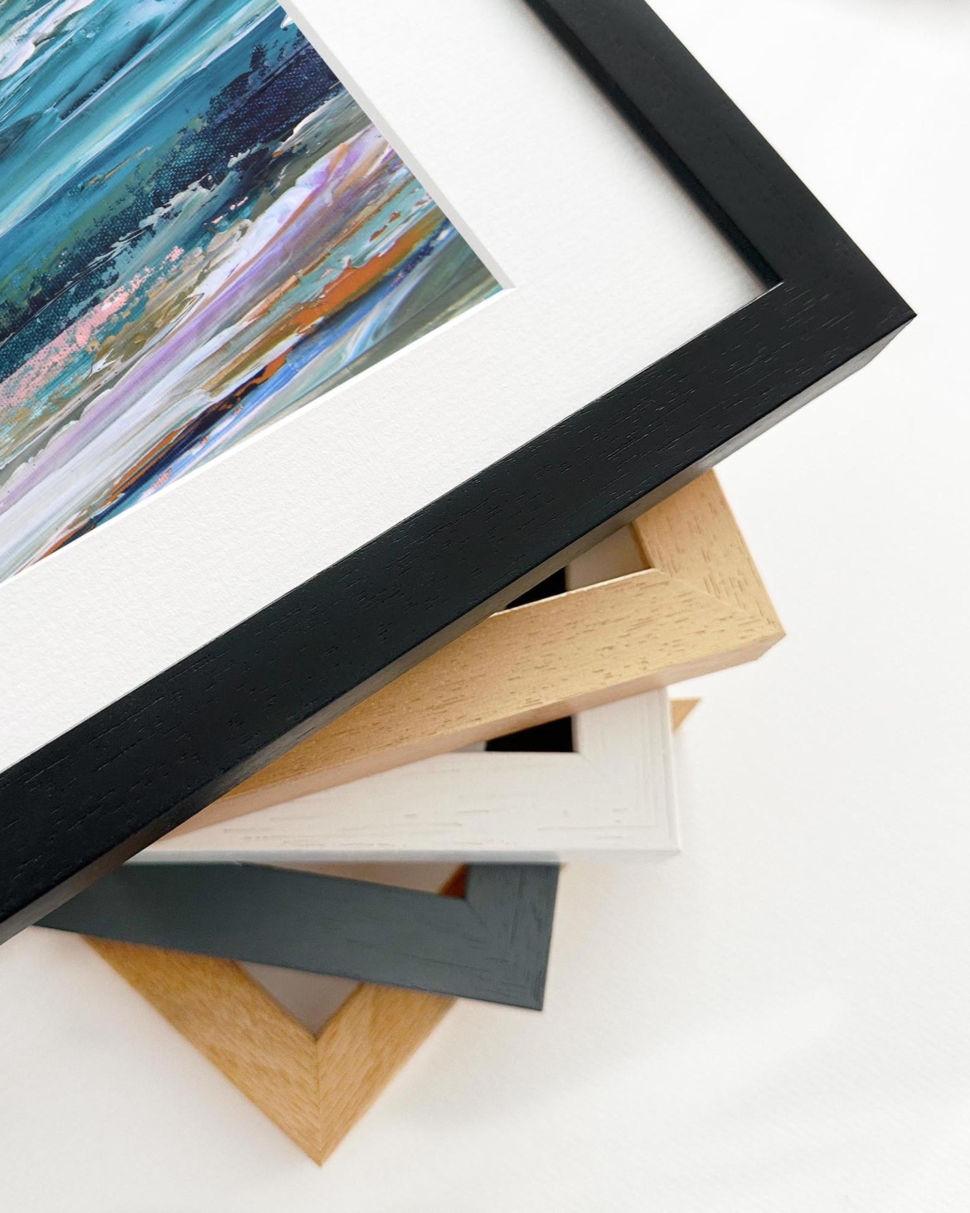 Stack of variously colored picture frames on a white surface with a framed artwork in the foreground.