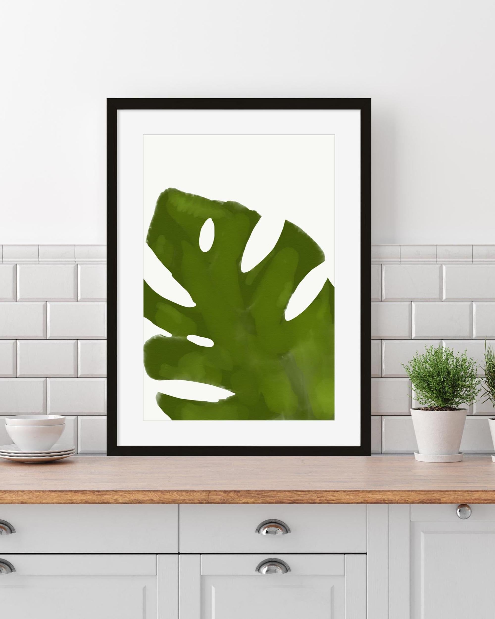 Framed green leaf print on a kitchen counter with white cabinets and plants.