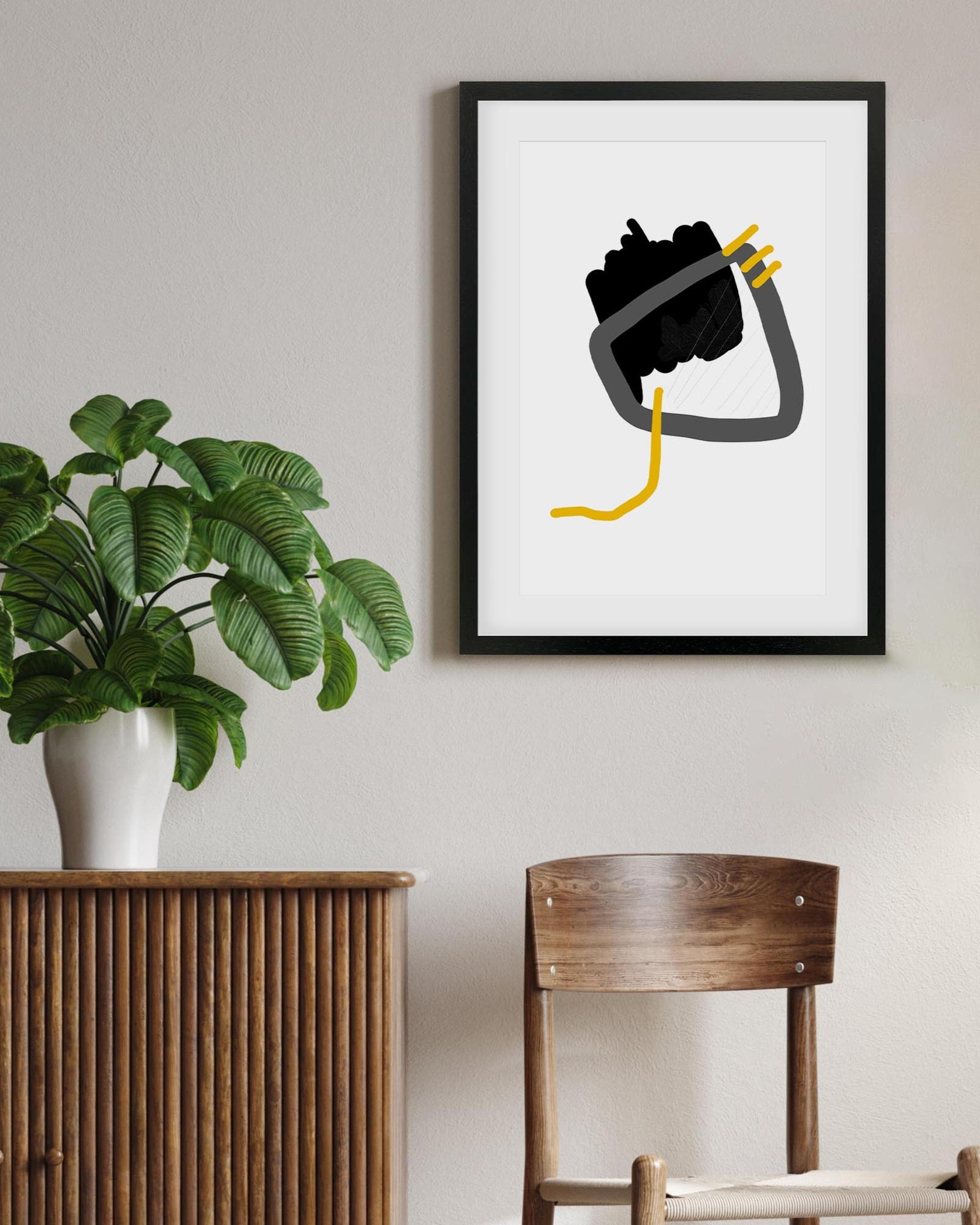 Framed abstract art piece on a wall with a plant and wooden furniture in the foreground.