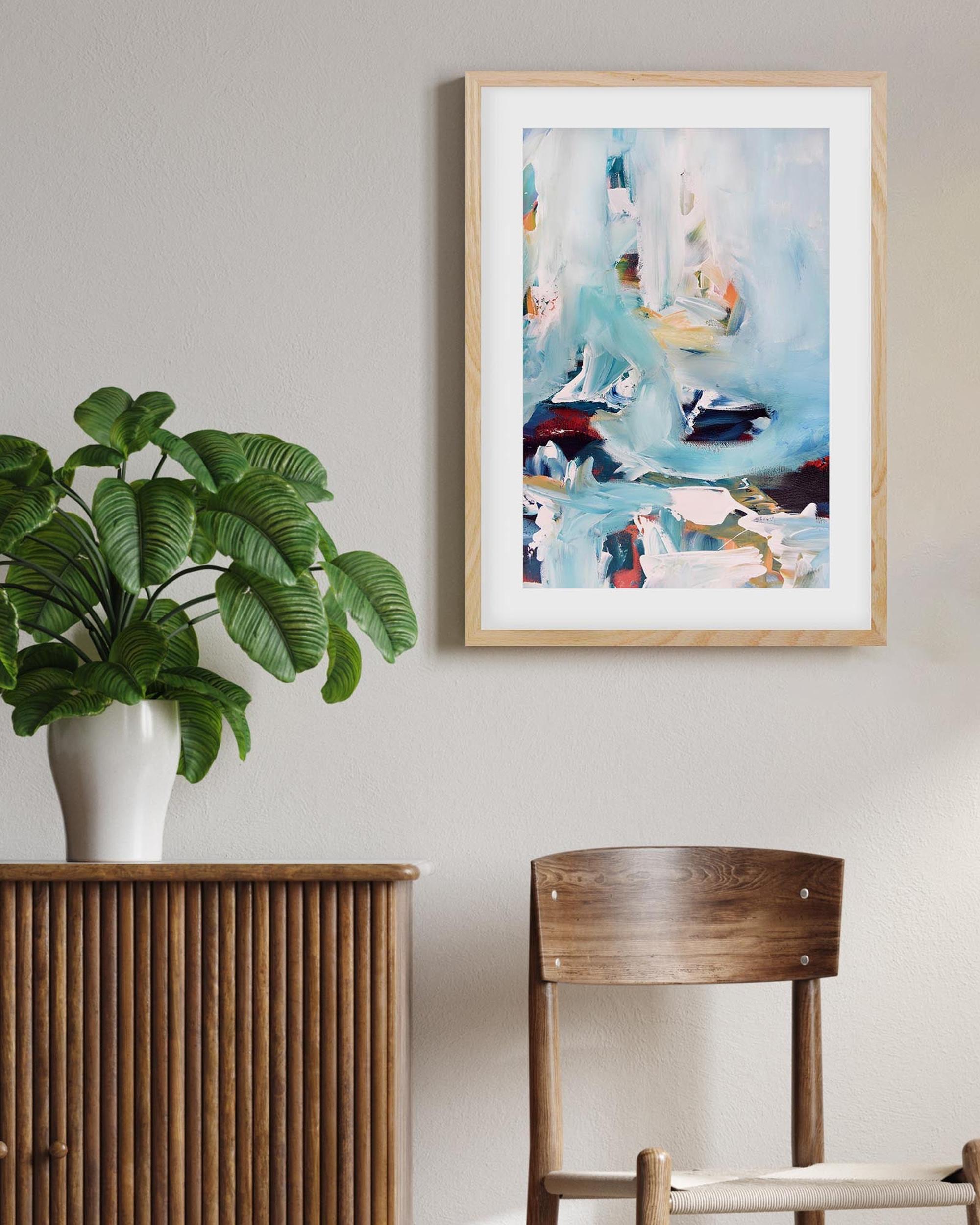 Framed abstract art piece on a wall above a wooden cabinet with a plant and chair.