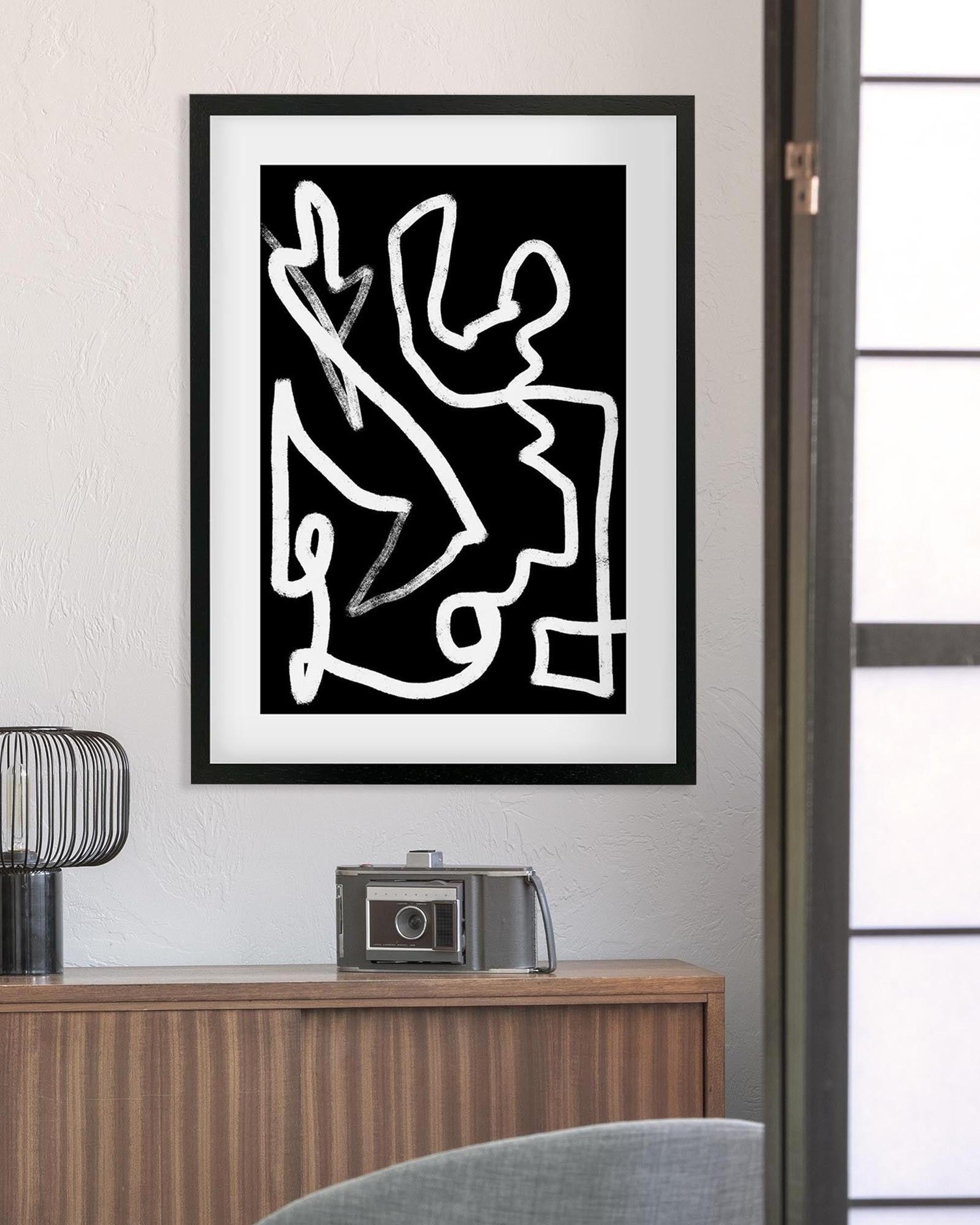 Framed abstract artwork on a wall above a wooden cabinet with a camera and decorative object.