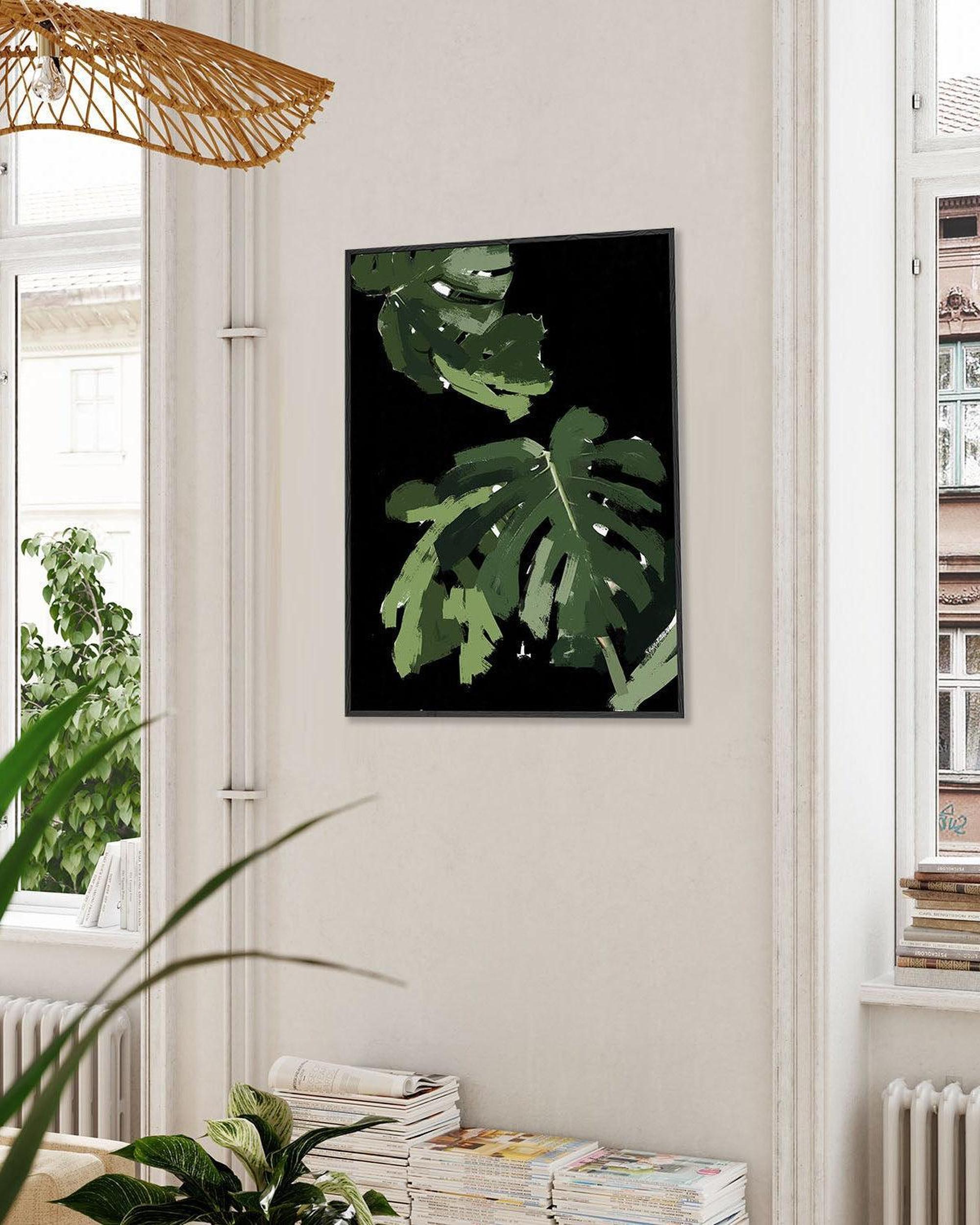 Framed artwork of green leaves on a wall in a room with plants and books.