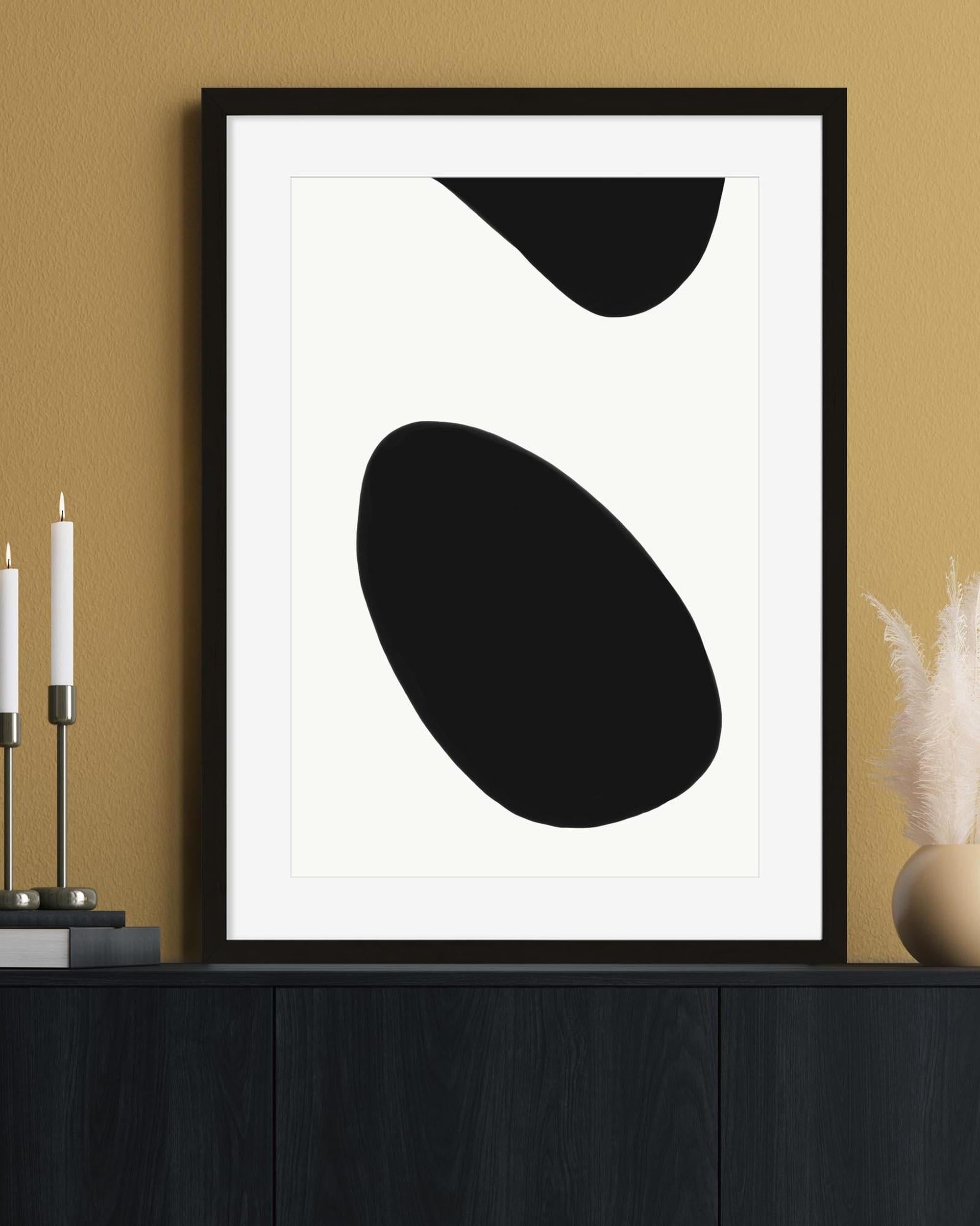 Framed abstract art print with black shapes on a white background, displayed on a wall.
