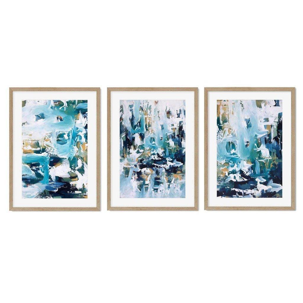 A set of three framed abstract art prints featuring teal and blue brushstrokes with yellow undertones displayed on a wall above a modern dining set.