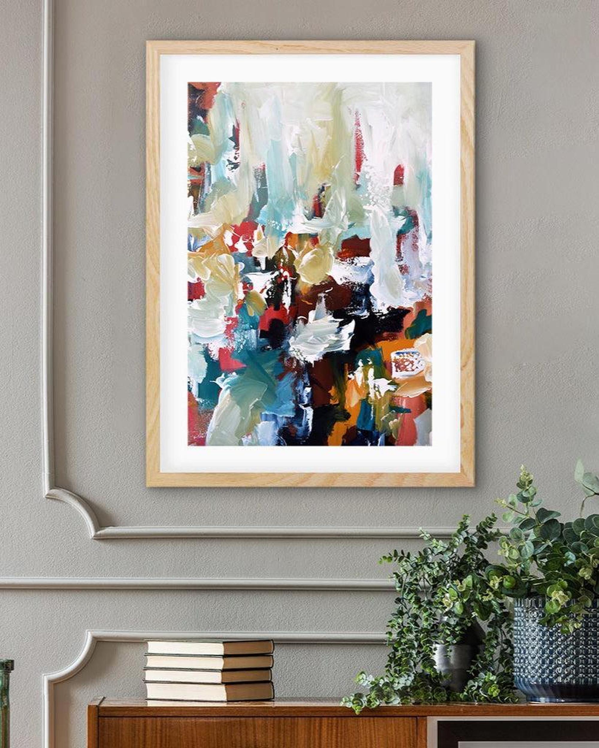 Framed abstract art piece on a wall with a plant and books below.