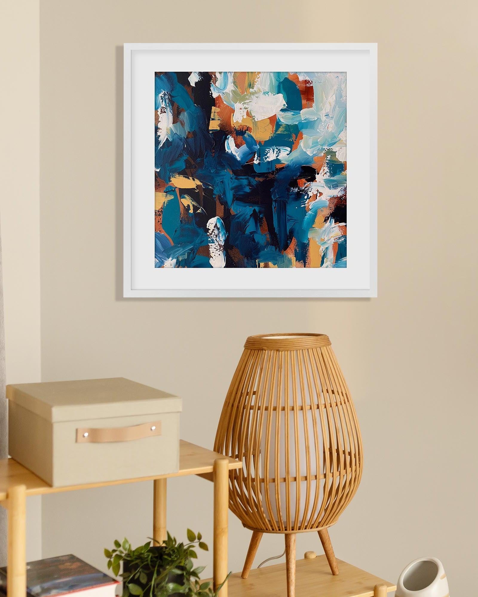 Framed abstract art piece on a wall above a wooden side table with a lamp and decorative box.