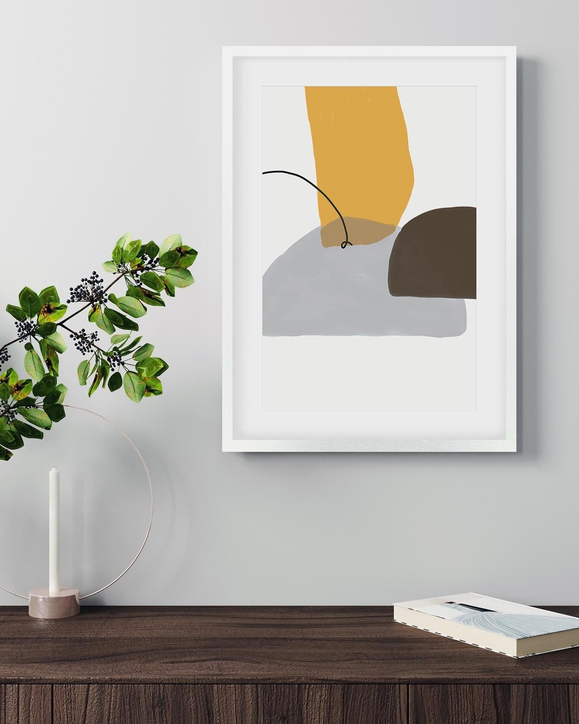 Abstract art print in a white frame on a wall above a wooden surface with a plant and book.