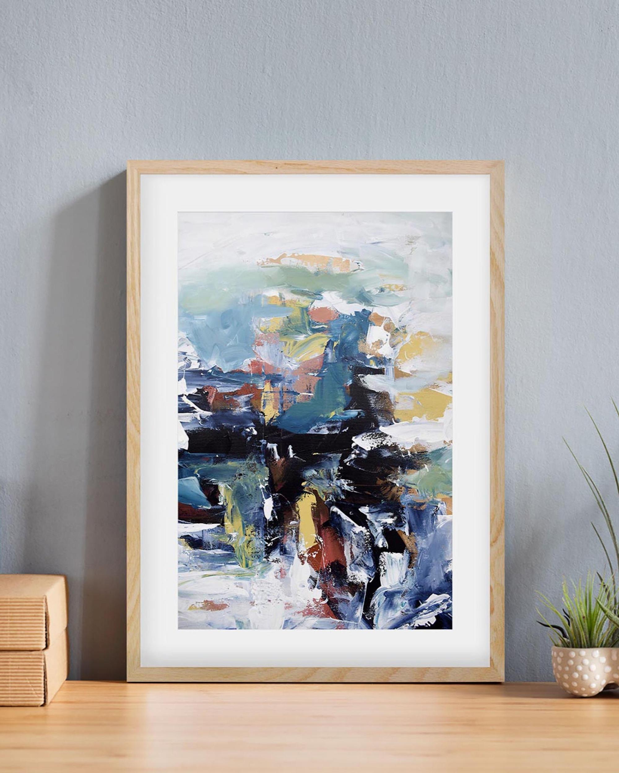 Framed abstract art print on a wall with a plant and wooden box in the foreground.