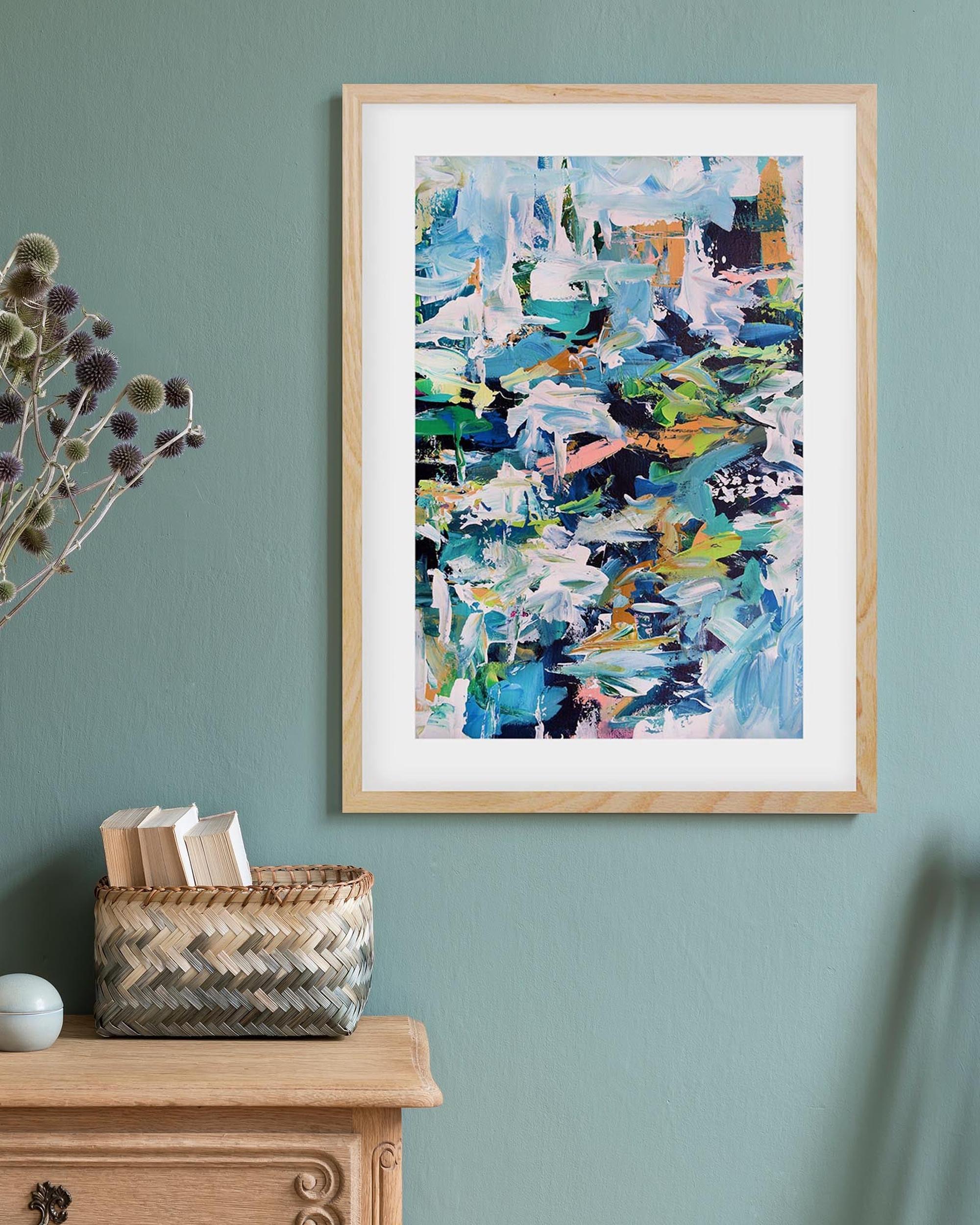 Framed abstract artwork on a teal wall with a wooden cabinet and basket.