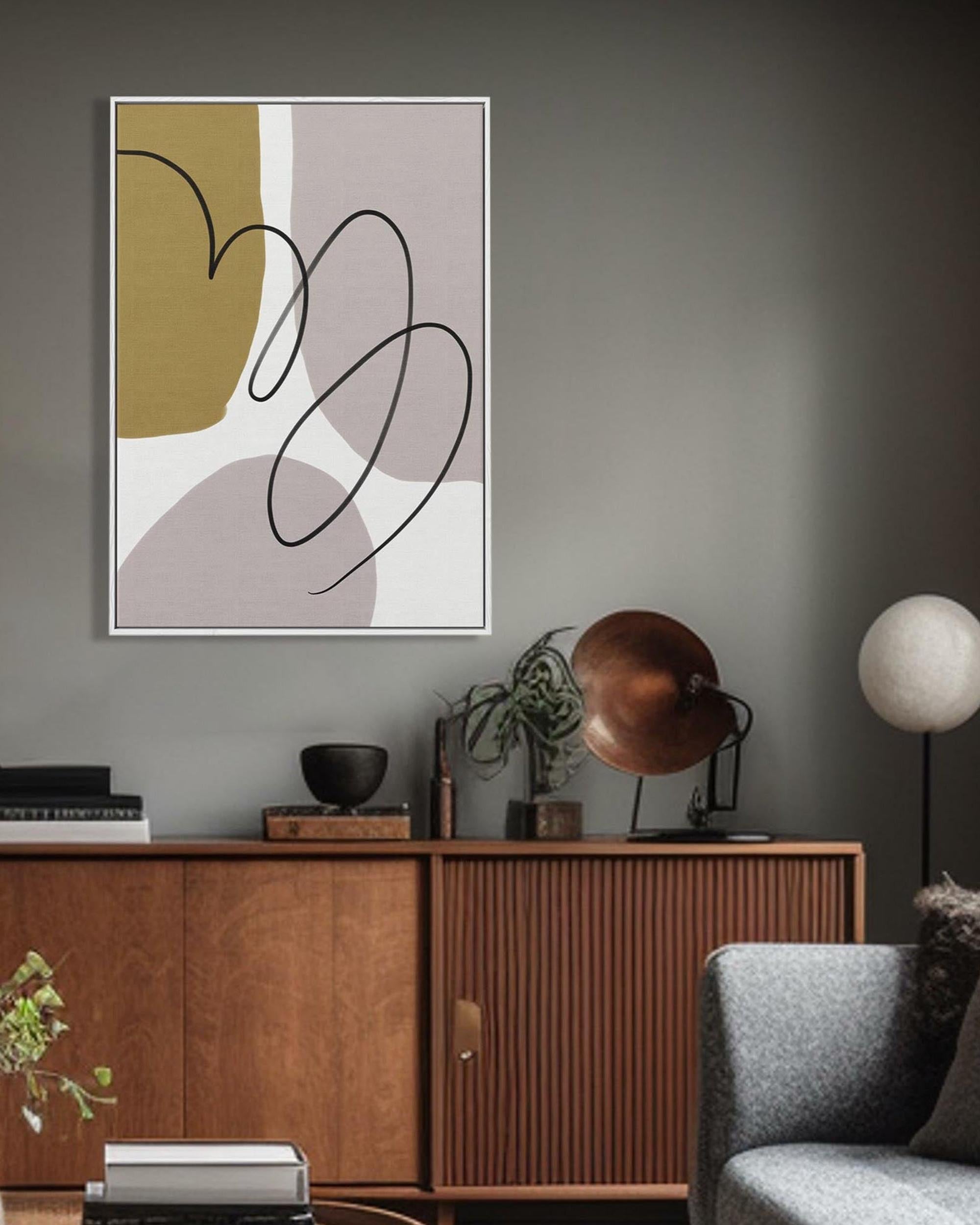 A framed canvas with abstract grey and mustard lines displayed above a wooden cabinet and beside a grey chair in a room with dark walls.