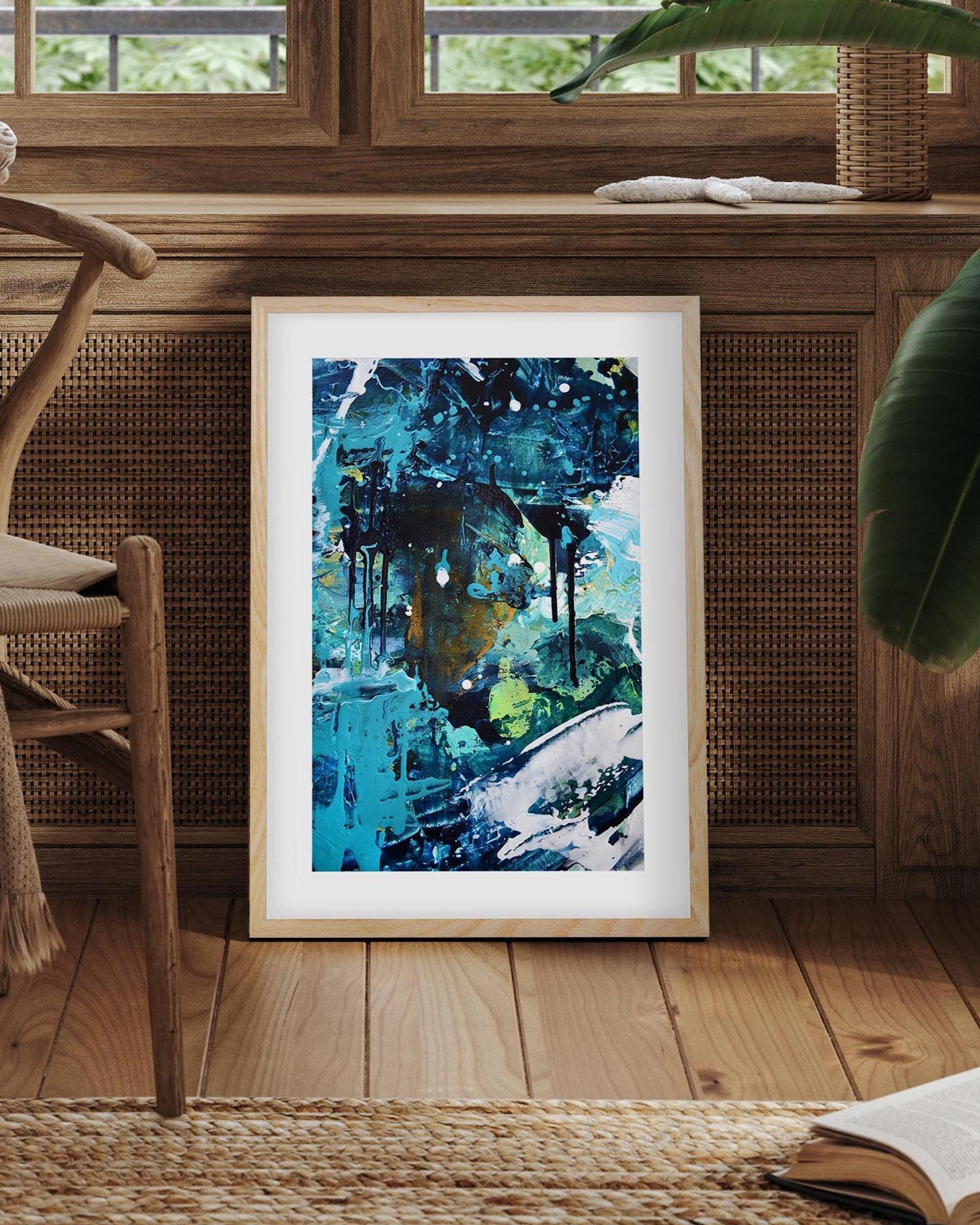 Framed abstract artwork on a wooden floor with a chair and books in the background