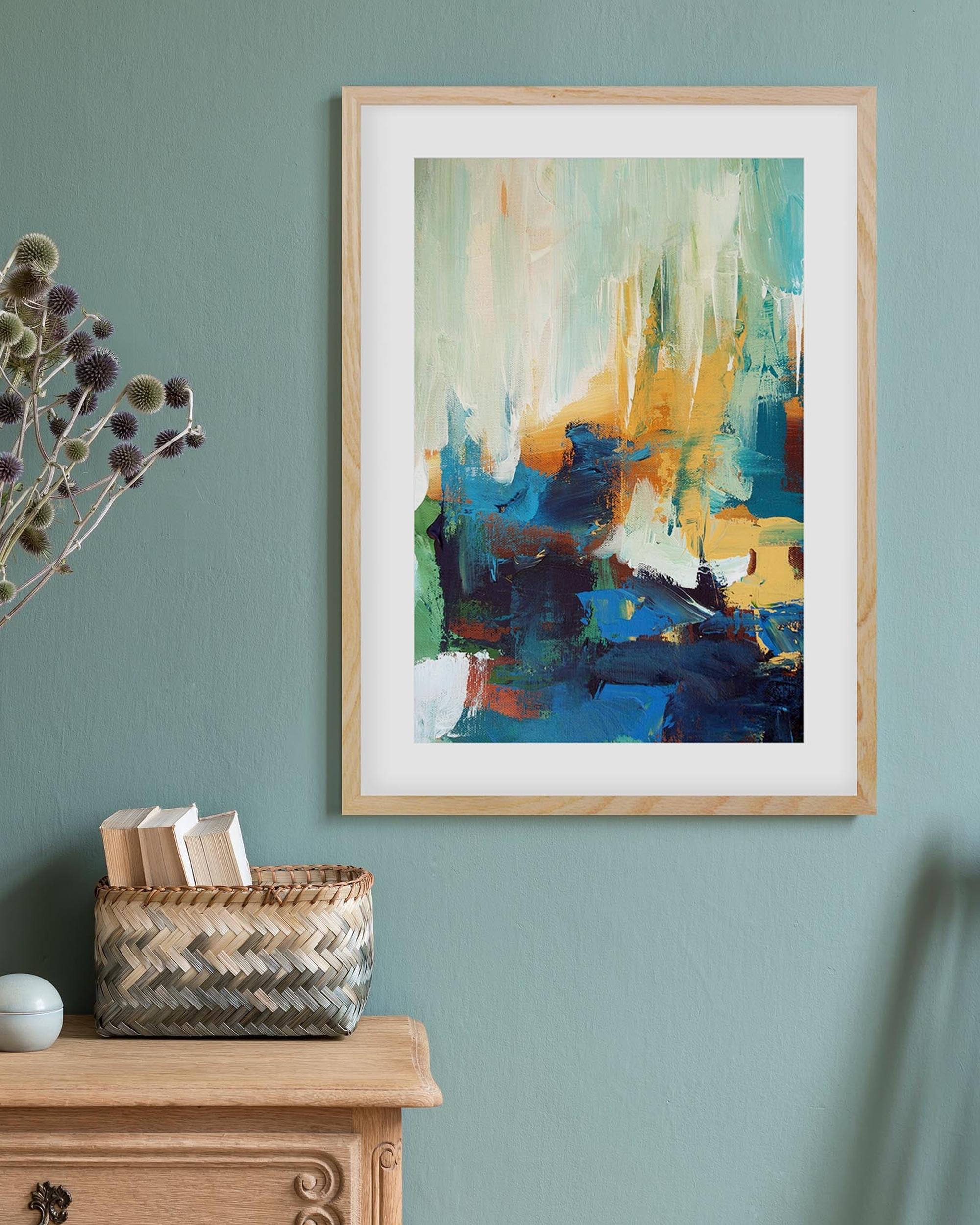 Framed abstract art on a wall with a vase of dried flowers and a basket below.