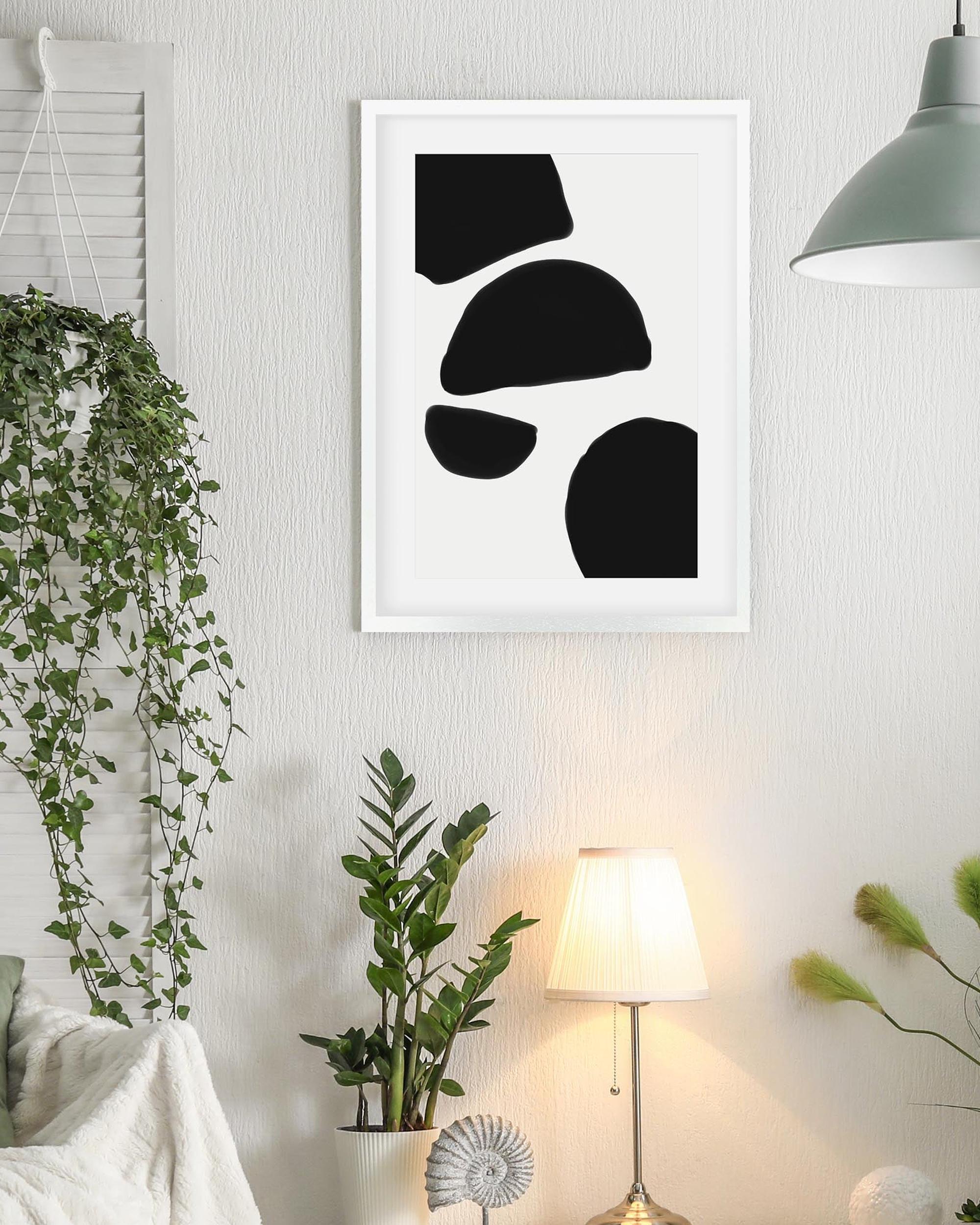 Framed abstract black and white artwork on a wall with plants and a lamp in the background.