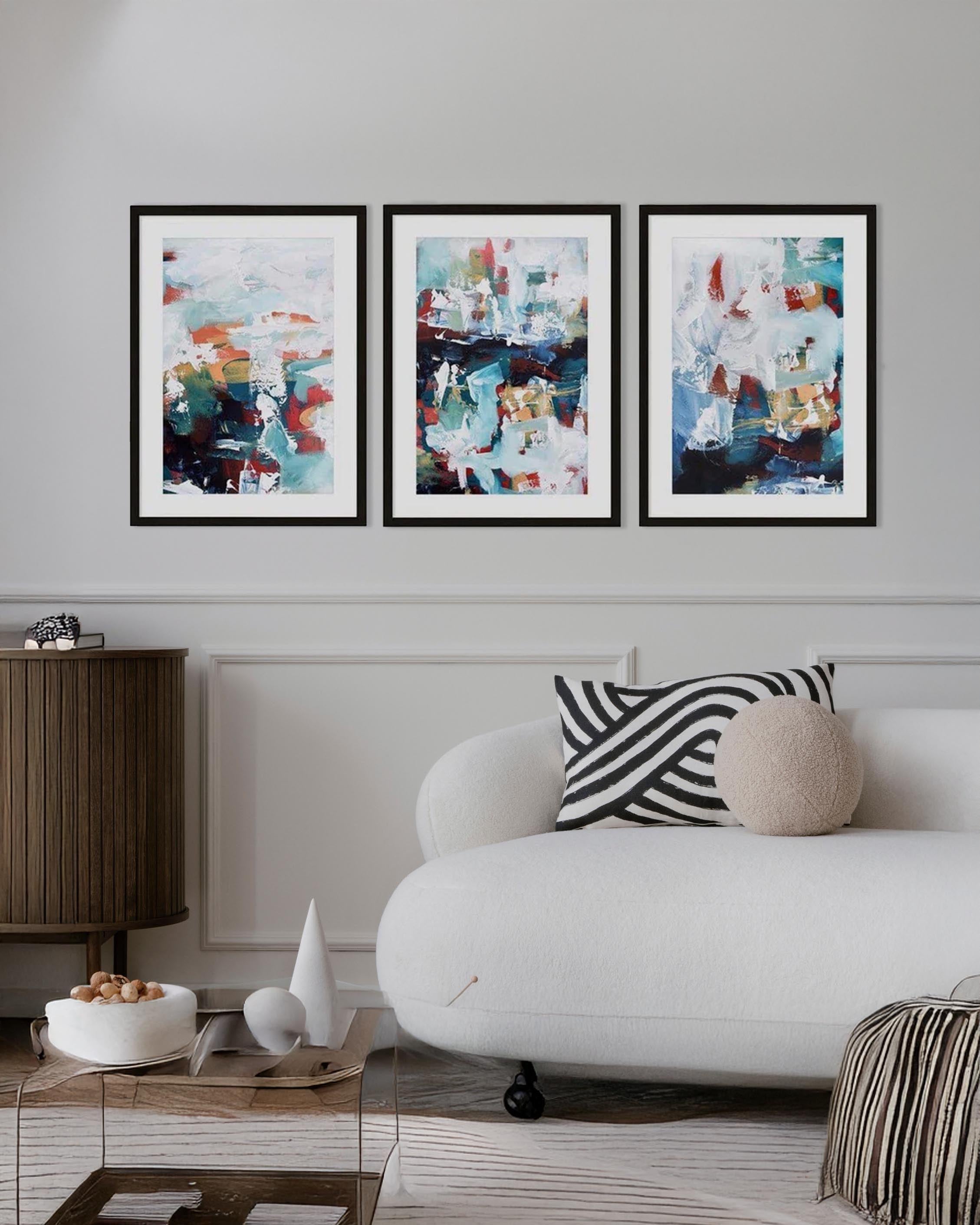 A set of three framed abstract art prints hanging on a wall over a white sofa, with a wooden side table and decorative objects in the foreground.