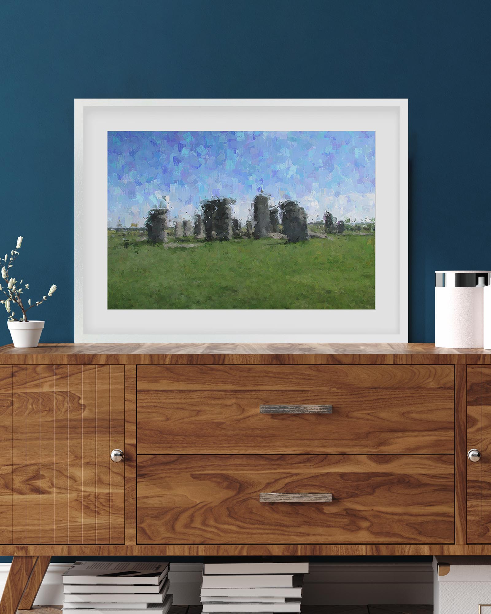 Framed artwork of stone structures in a field on a wooden cabinet against a blue wall.