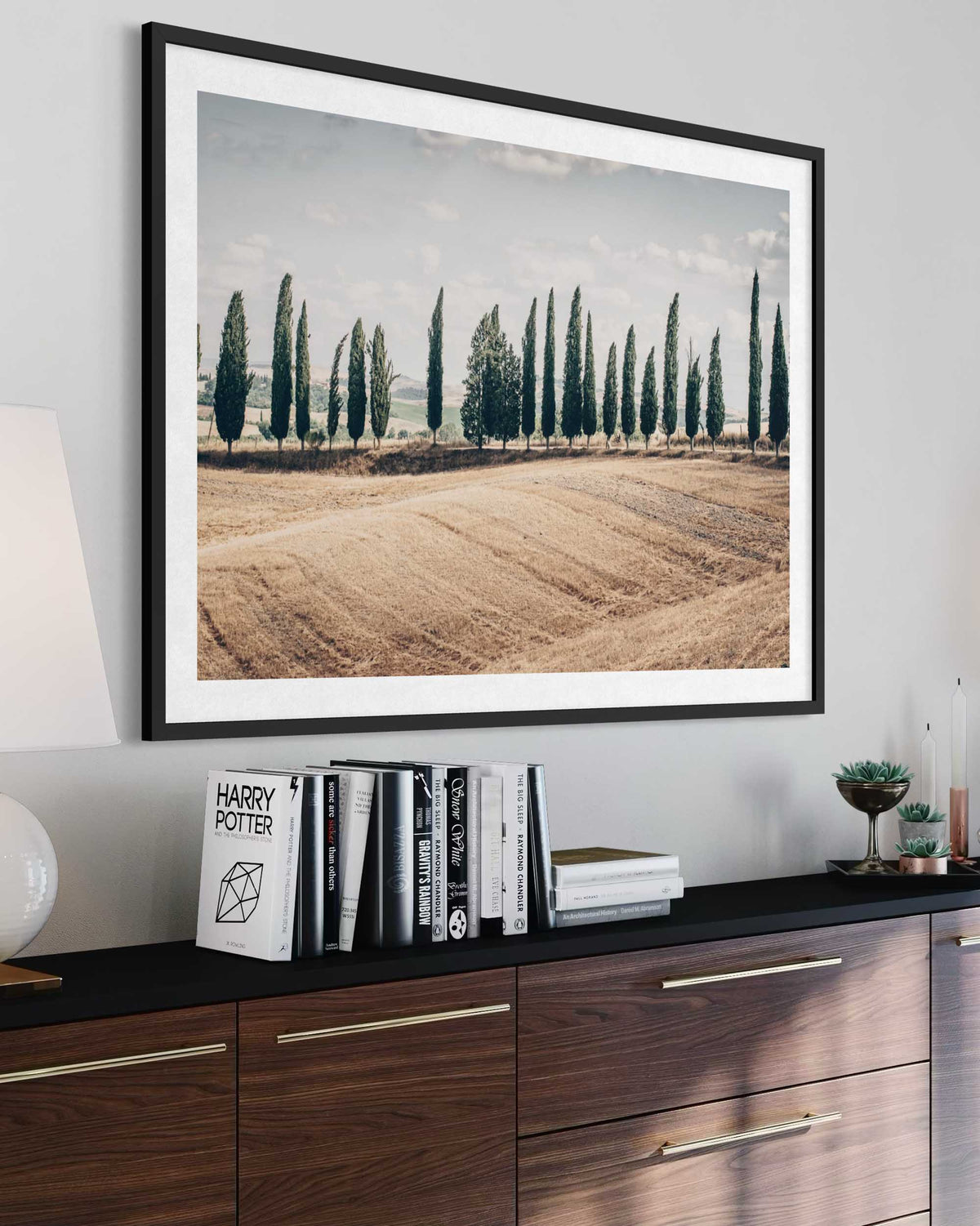 Framed landscape print of a field with trees on a wall above a wooden cabinet.