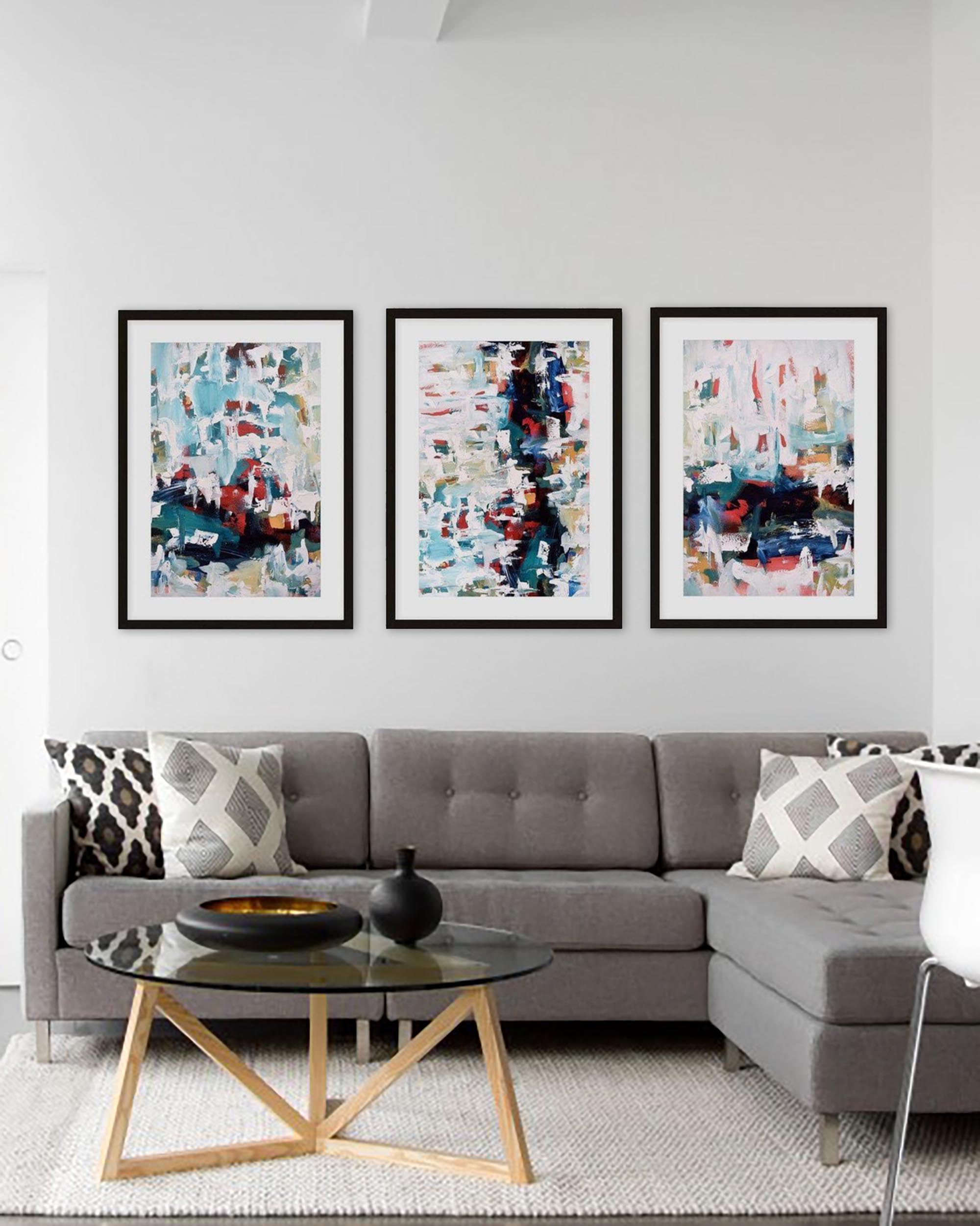 A set of three framed abstract art prints displayed on a wall above a gray sofa with patterned cushions.