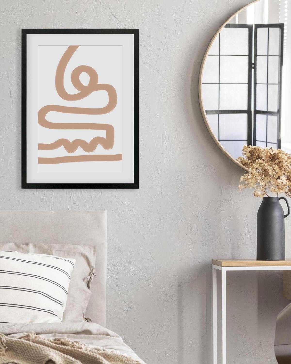 Framed abstract art piece on a wall above a bed with a round mirror and vase in the background.