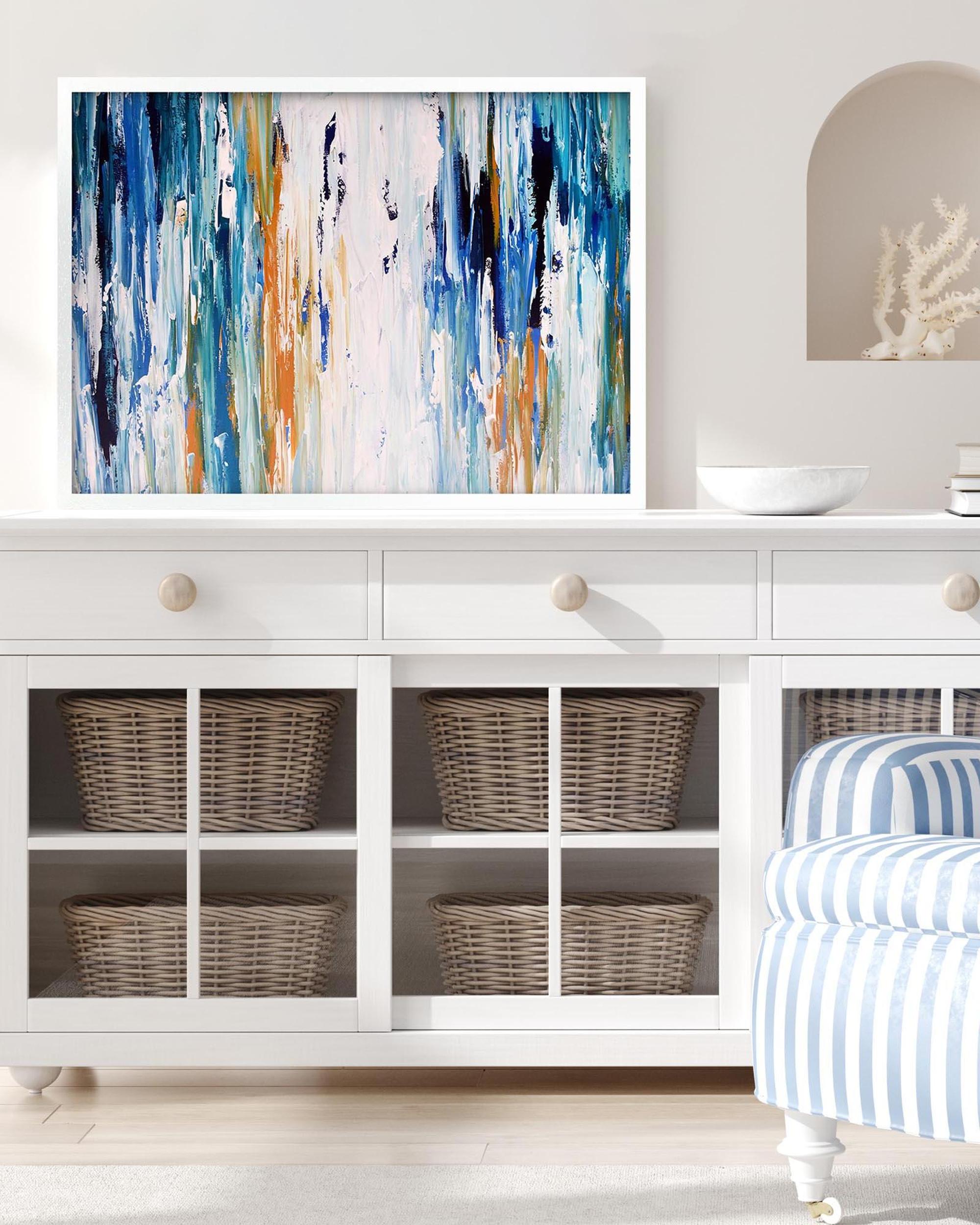 Framed abstract painting on a white wall above a white cabinet with wicker baskets.