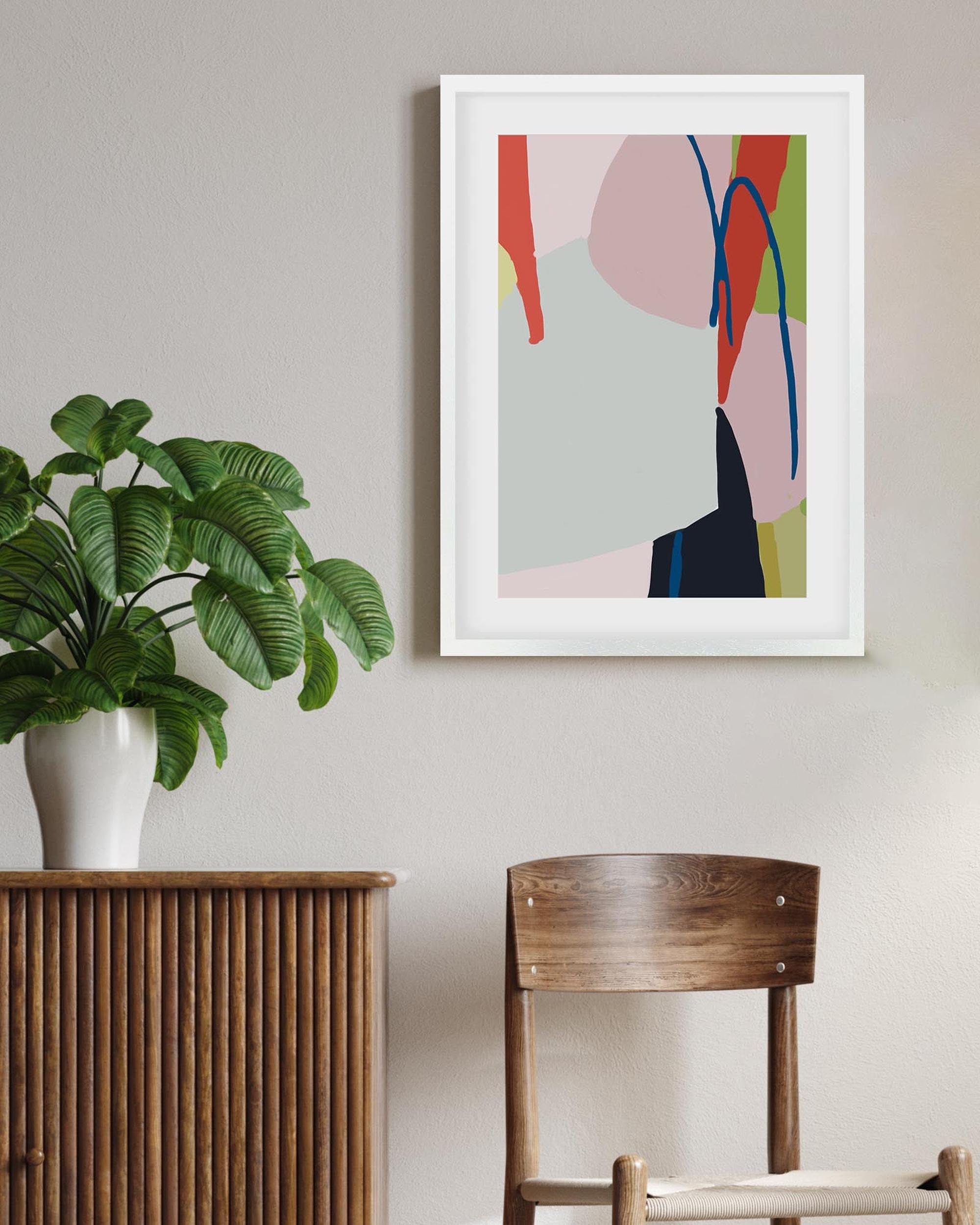 Framed abstract art on a wall with a plant and wooden chair below.
