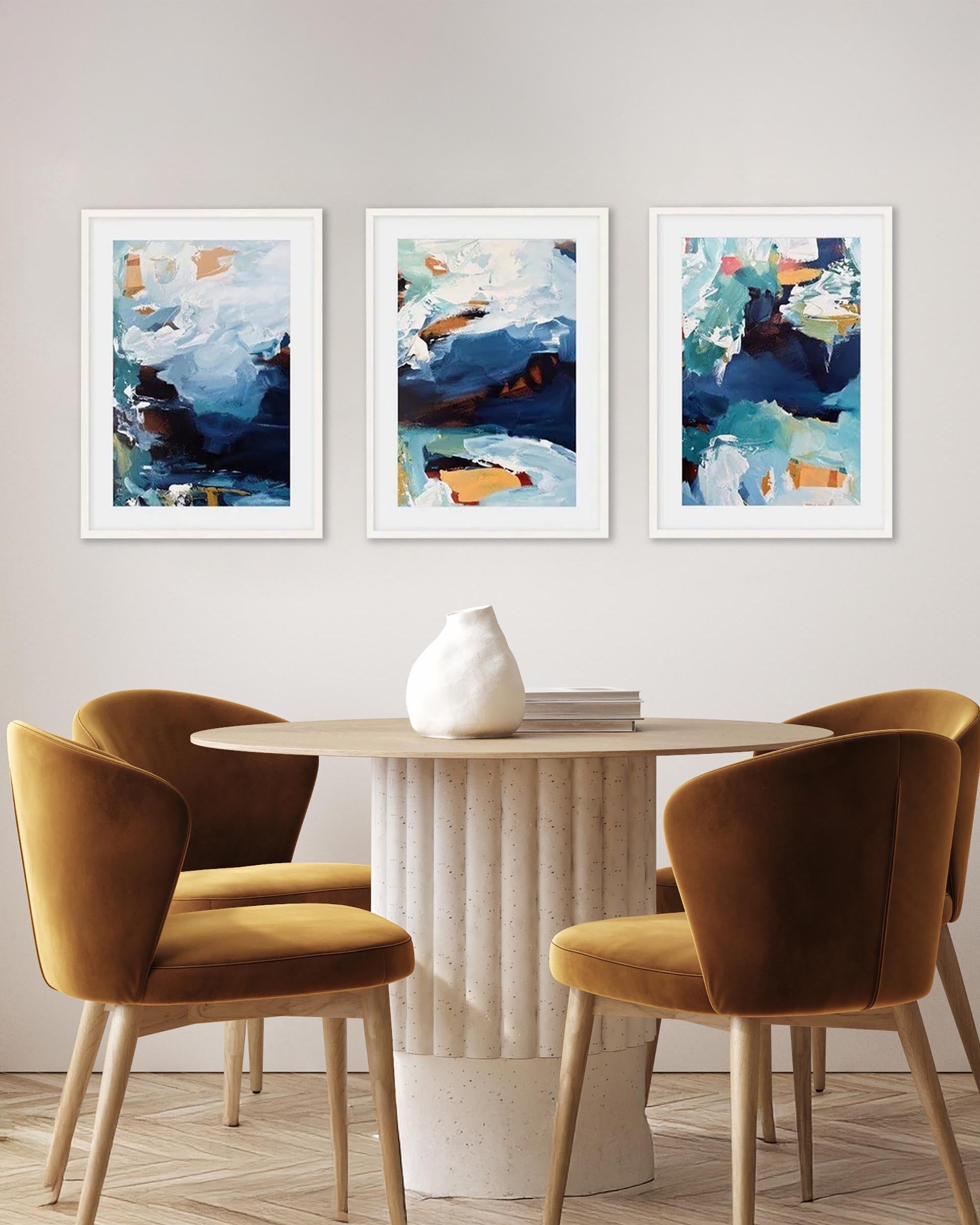 A set of three framed abstract art prints displayed on a wall above a white round table with two chairs in a room.