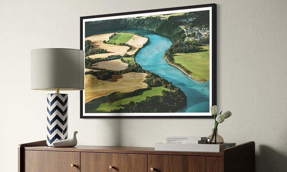 Framed landscape print of a river and fields on a wall above a wooden sideboard with decor items.