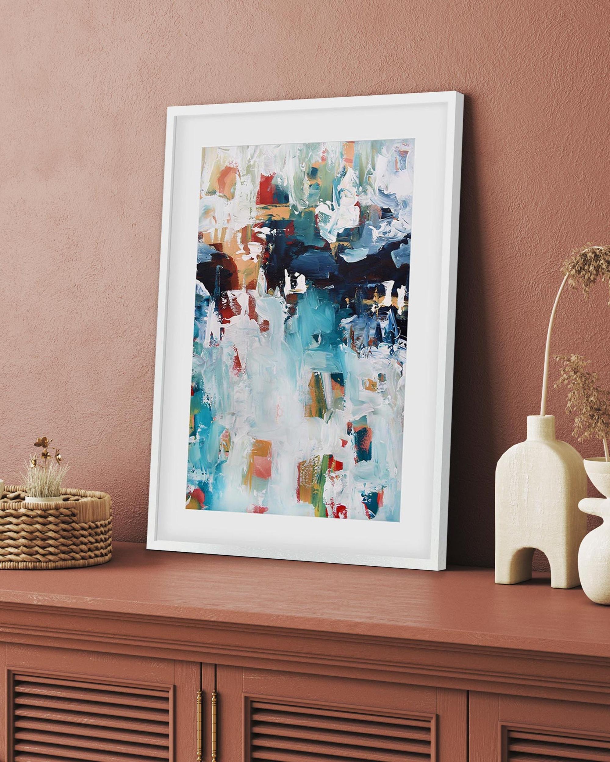 Framed abstract art on a wooden surface with decorative items against a pink wall.