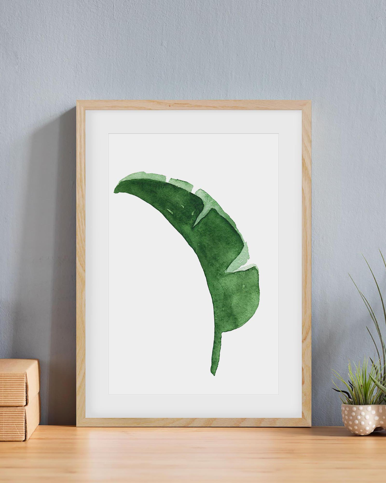 Framed artwork of a green leaf on a white background, displayed on a wooden surface with a light gray wall.