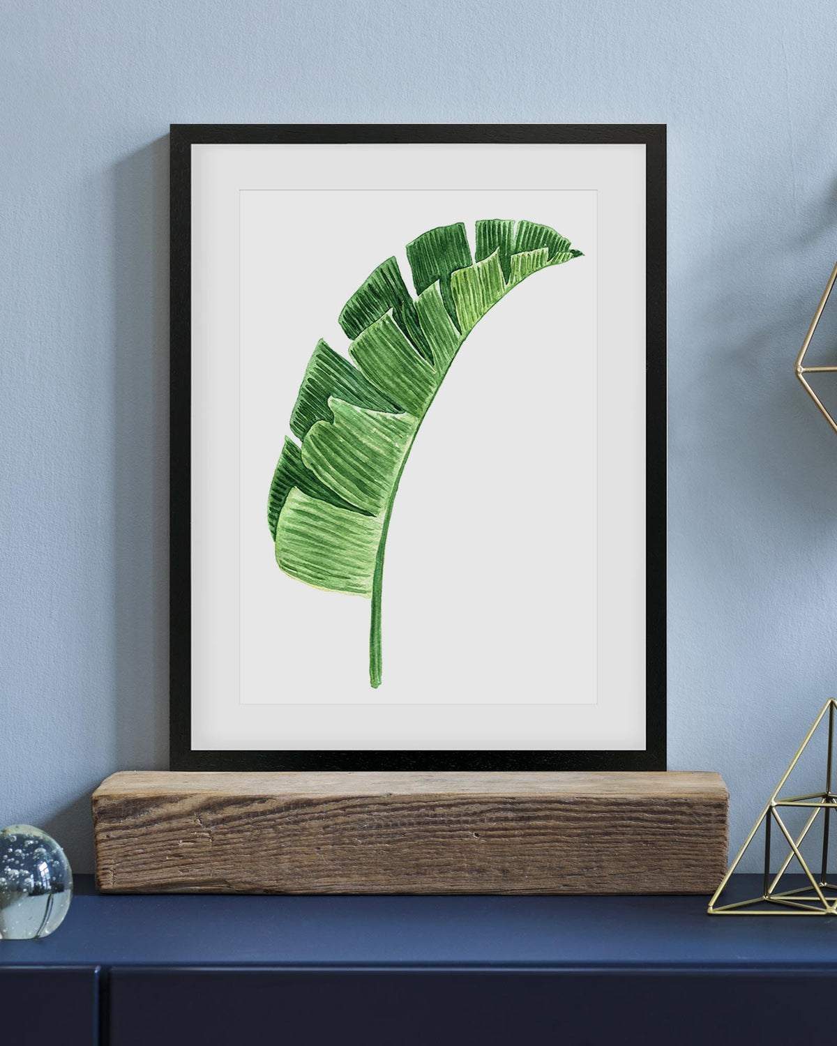 Framed artwork of a green leaf on a white background, displayed on a shelf against a blue wall.