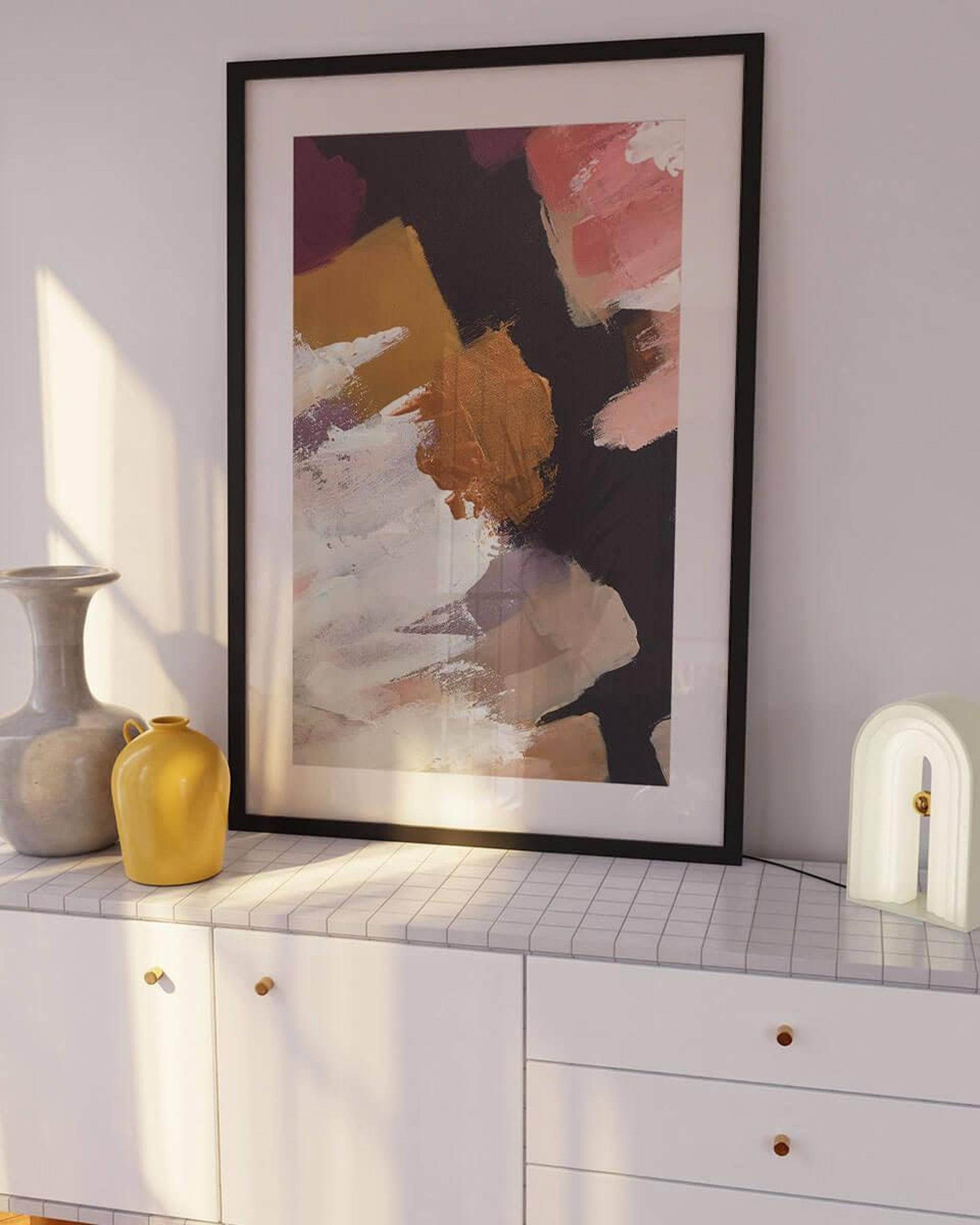 Framed abstract art on a wall above a white cabinet with decorative items.