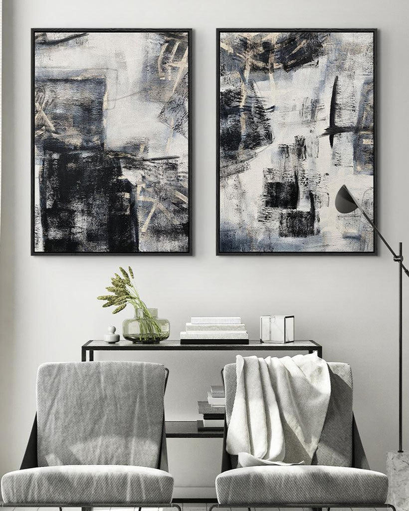 A set of two contemporary abstract art canvases with grey tones and light gold undertones, displayed above a white console table in a room with wooden chairs and a window.