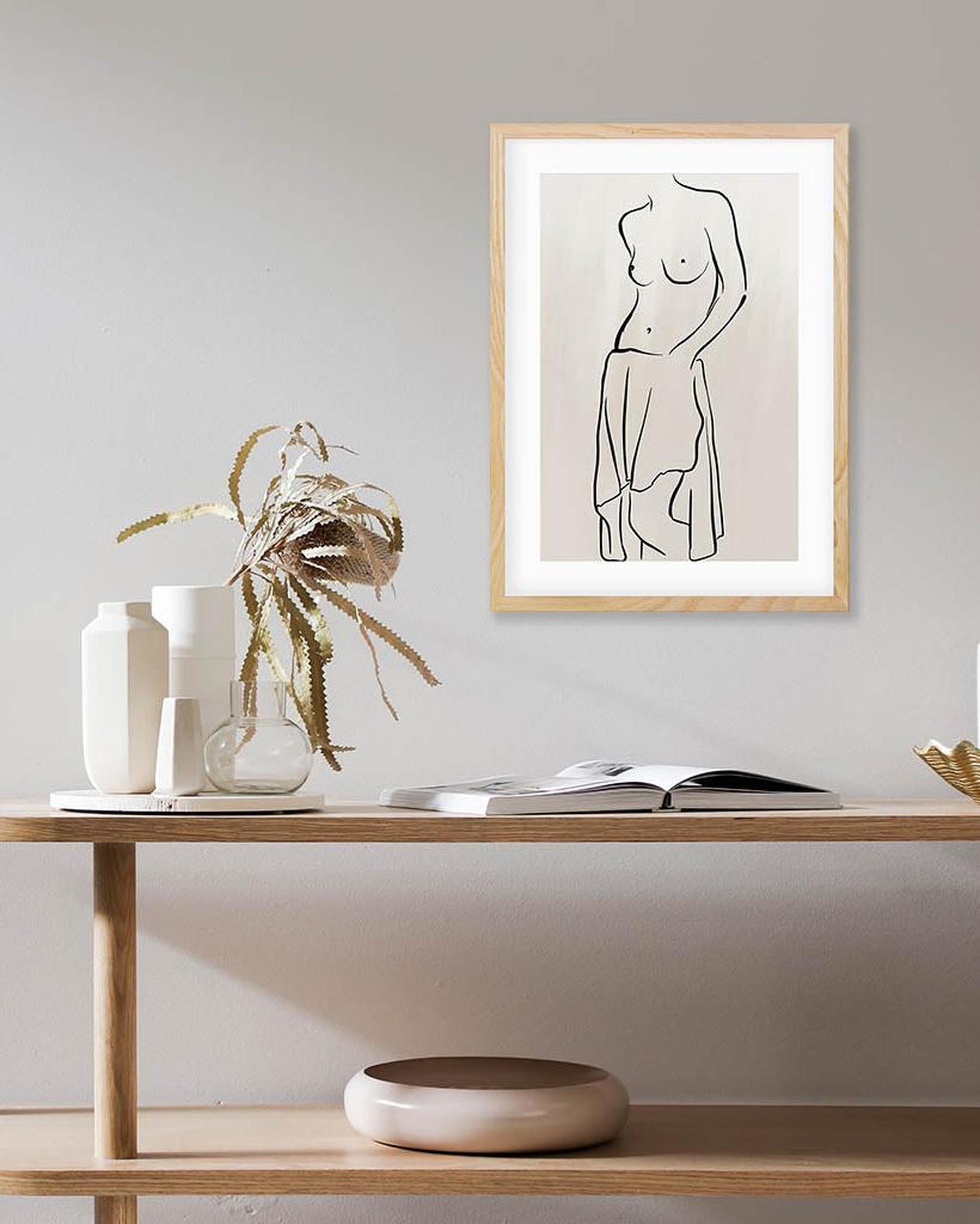 Framed abstract line art of a nude figure on a wall above a wooden shelf with decor items.