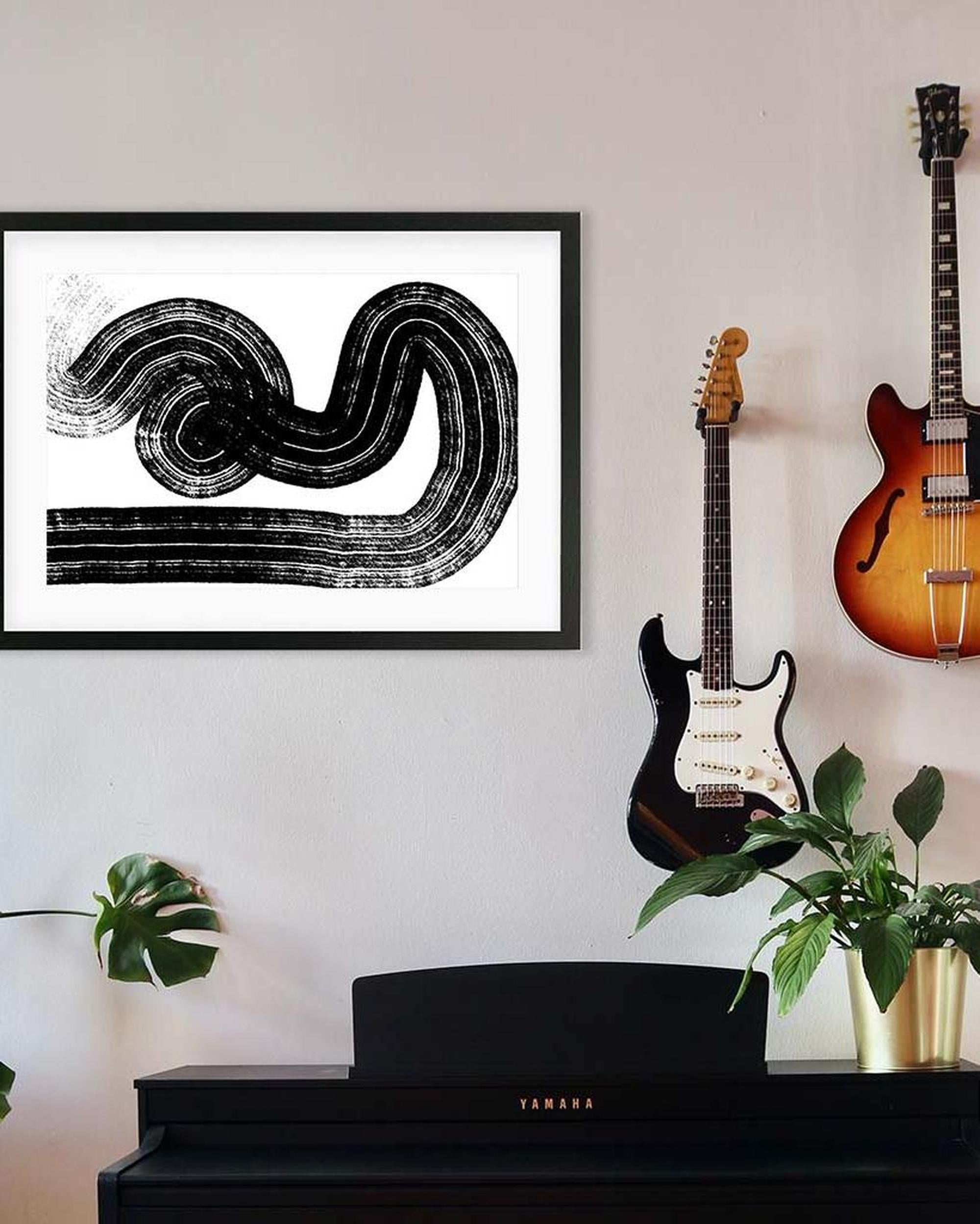 Guitar and bass on a wall with framed abstract art above a Yamaha piano.