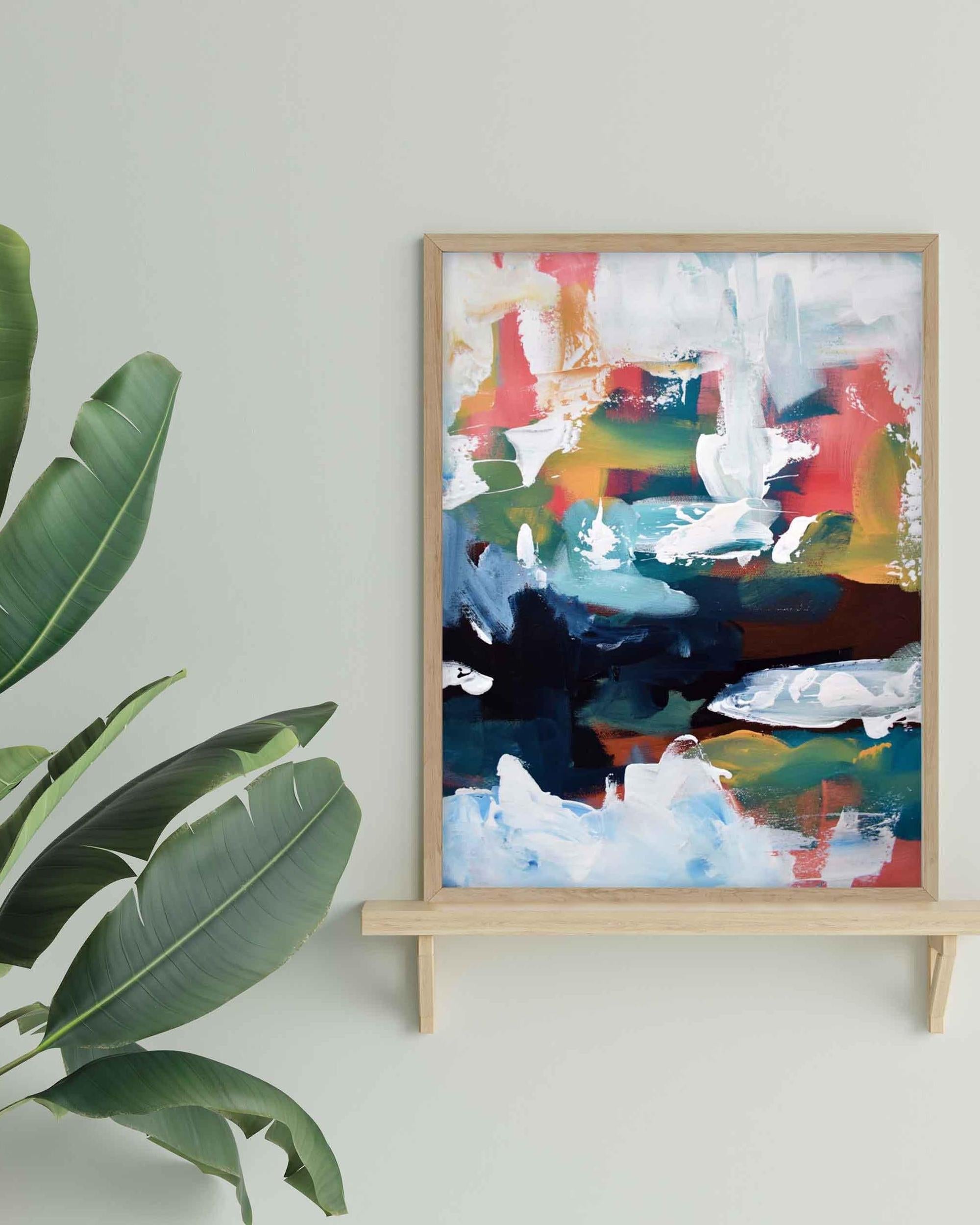 Abstract painting in a wooden frame on a stand against a light wall with a plant to the left.