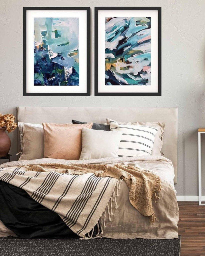 Two abstract art pieces on a wall above a bed with pillows and a blanket.