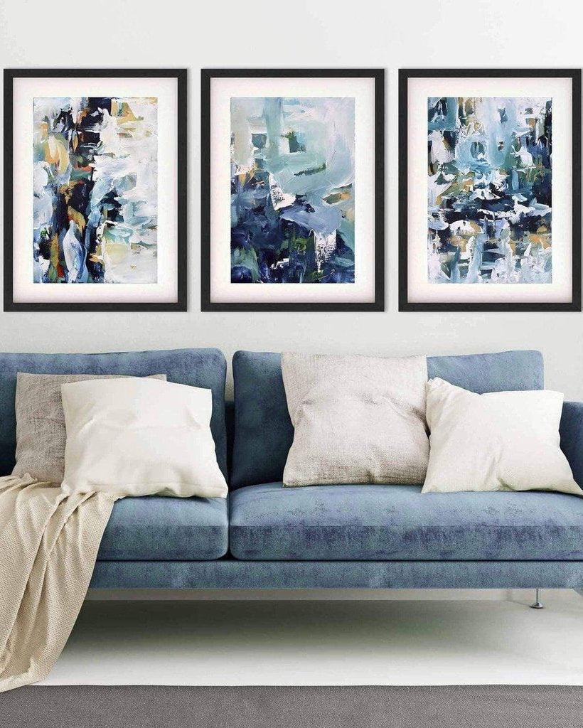 A set of three framed abstract blue art prints displayed above a blue sofa with a plant and wooden side table in a room.