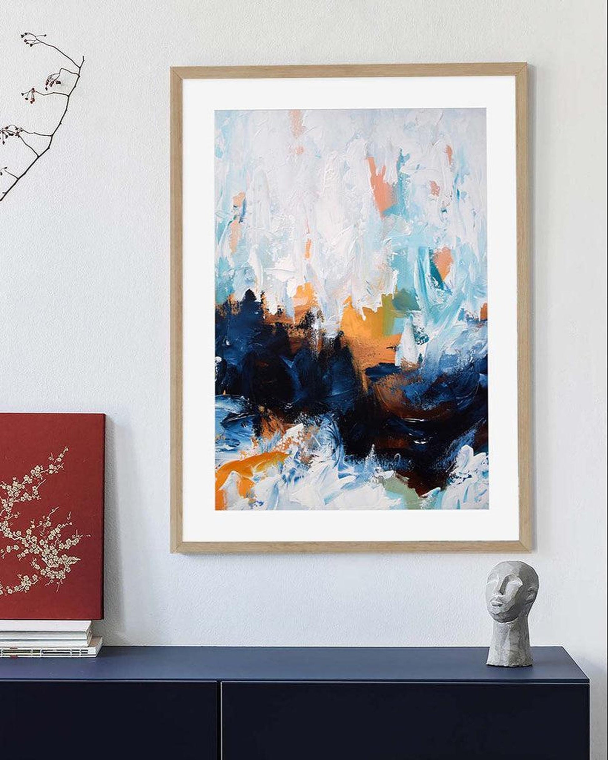 Abstract art print in a wooden frame on a wall above a dark surface with a red book and a gray sculpture.