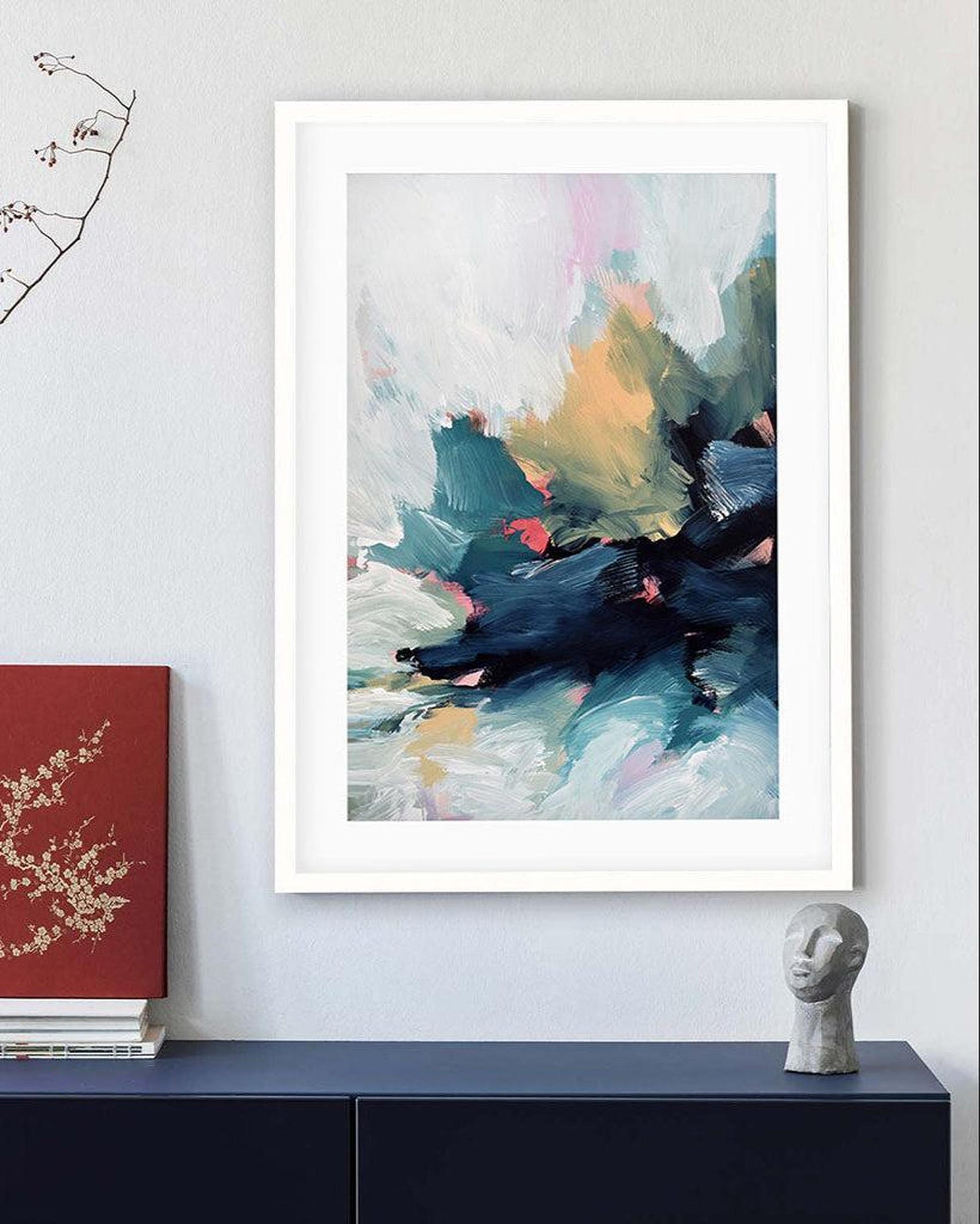 Abstract art print in a white frame on a wall above a dark surface with a red book and gray sculpture.
