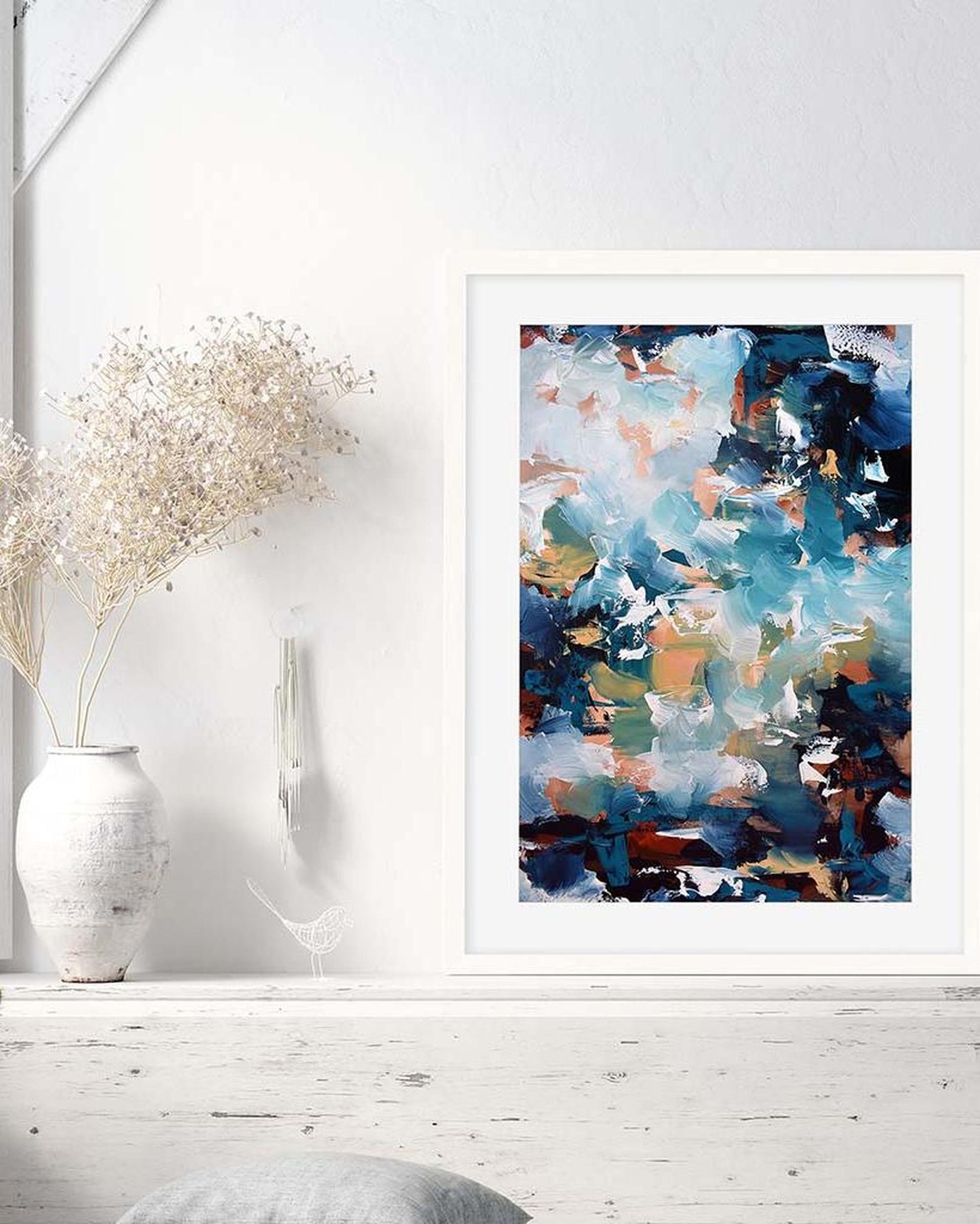Abstract painting in a white frame on a wall with a vase and decorative items.