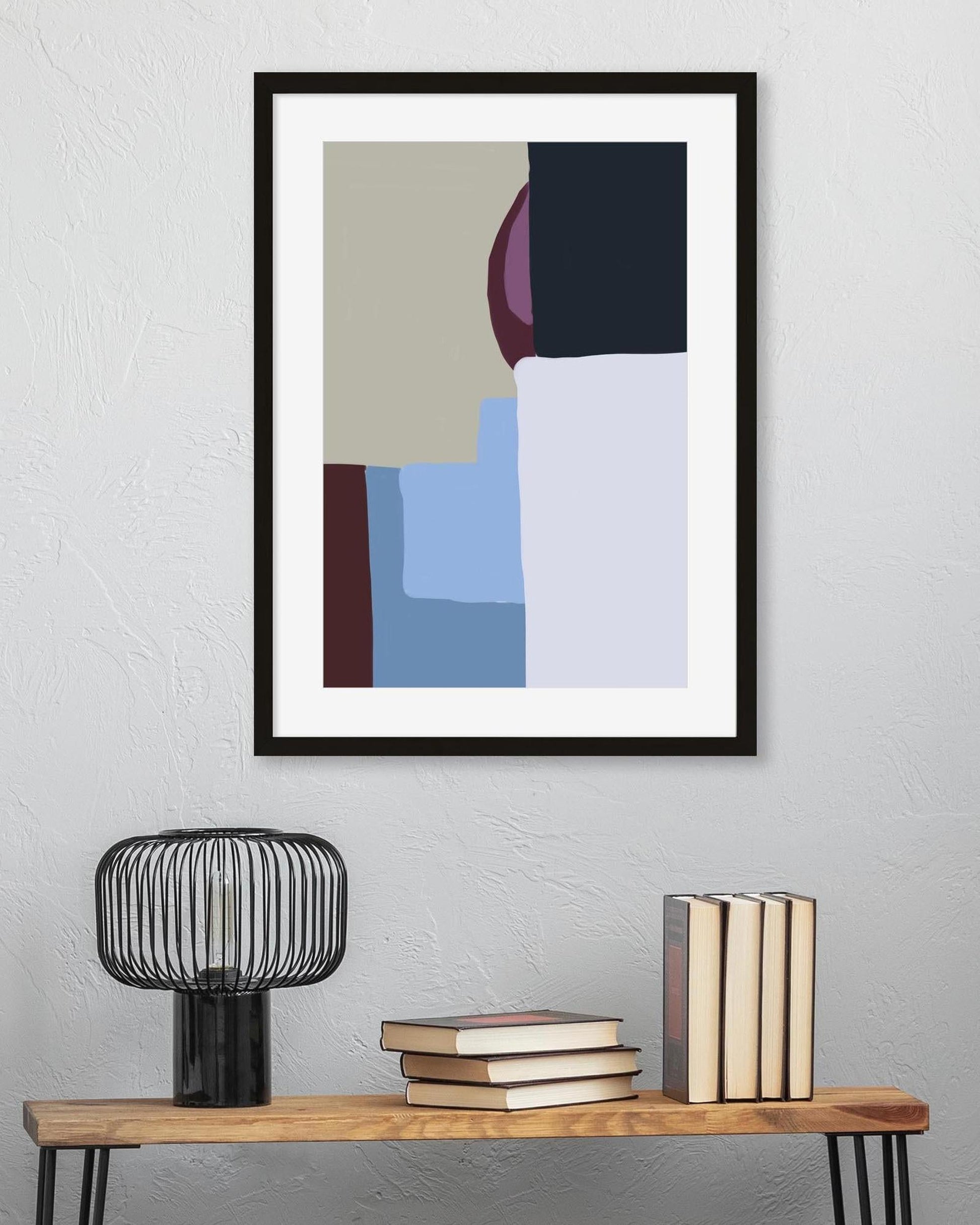 Framed abstract art piece on a wall above a wooden table with books and a lamp.