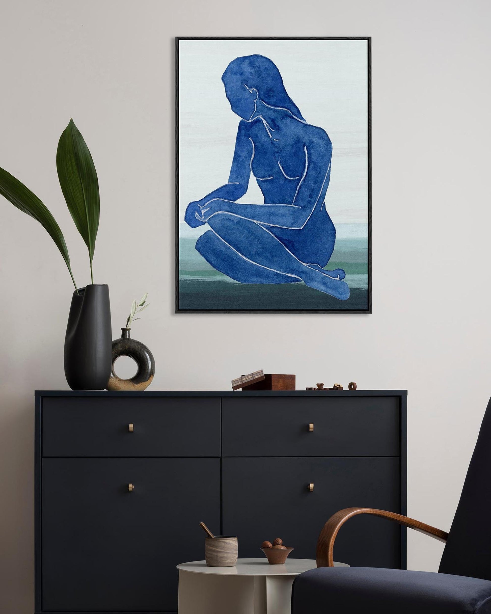 Framed abstract art of a blue figure on a wall above a black dresser.