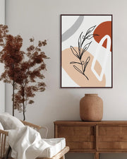 A framed canvas with abstract botanical shapes displayed above a wooden cabinet in a room with a chair and a decorative plant.