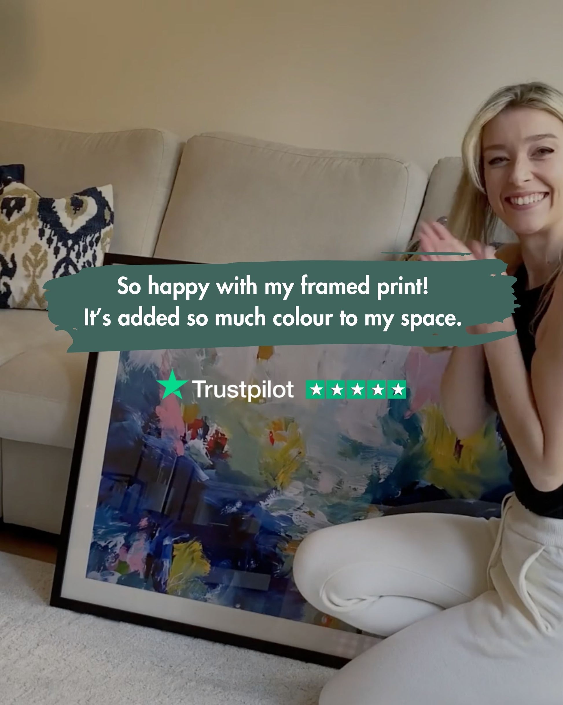 Woman sitting on a couch with a framed colorful print, smiling and holding a Trustpilot review card.