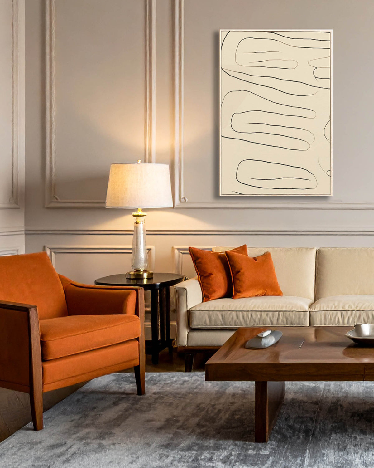 A framed abstract line art print with black lines on a cream background hangs on a gray paneled wall in a modern living room setting with an orange armchair, a beige sofa with orange pillows, and a wooden coffee table.