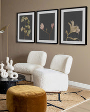 Modern living room with white textured chairs, brown ottoman, and floral wall art featuring three botanical wall art prints on a grey background.