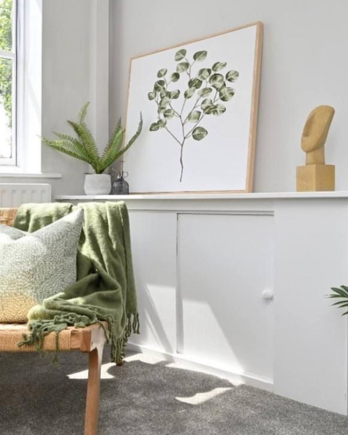 Nursery room with a wooden bench, green blanket, and framed plant art on a white wall.