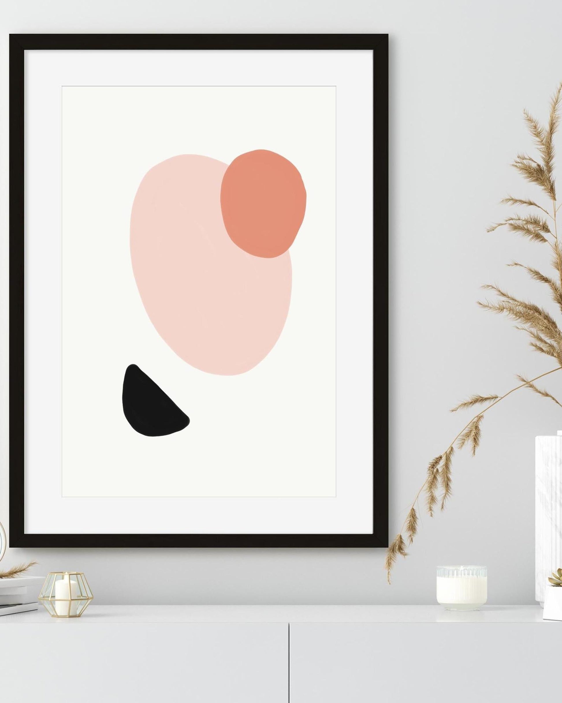 Framed abstract art print with pink, orange, and black shapes on a white wall.